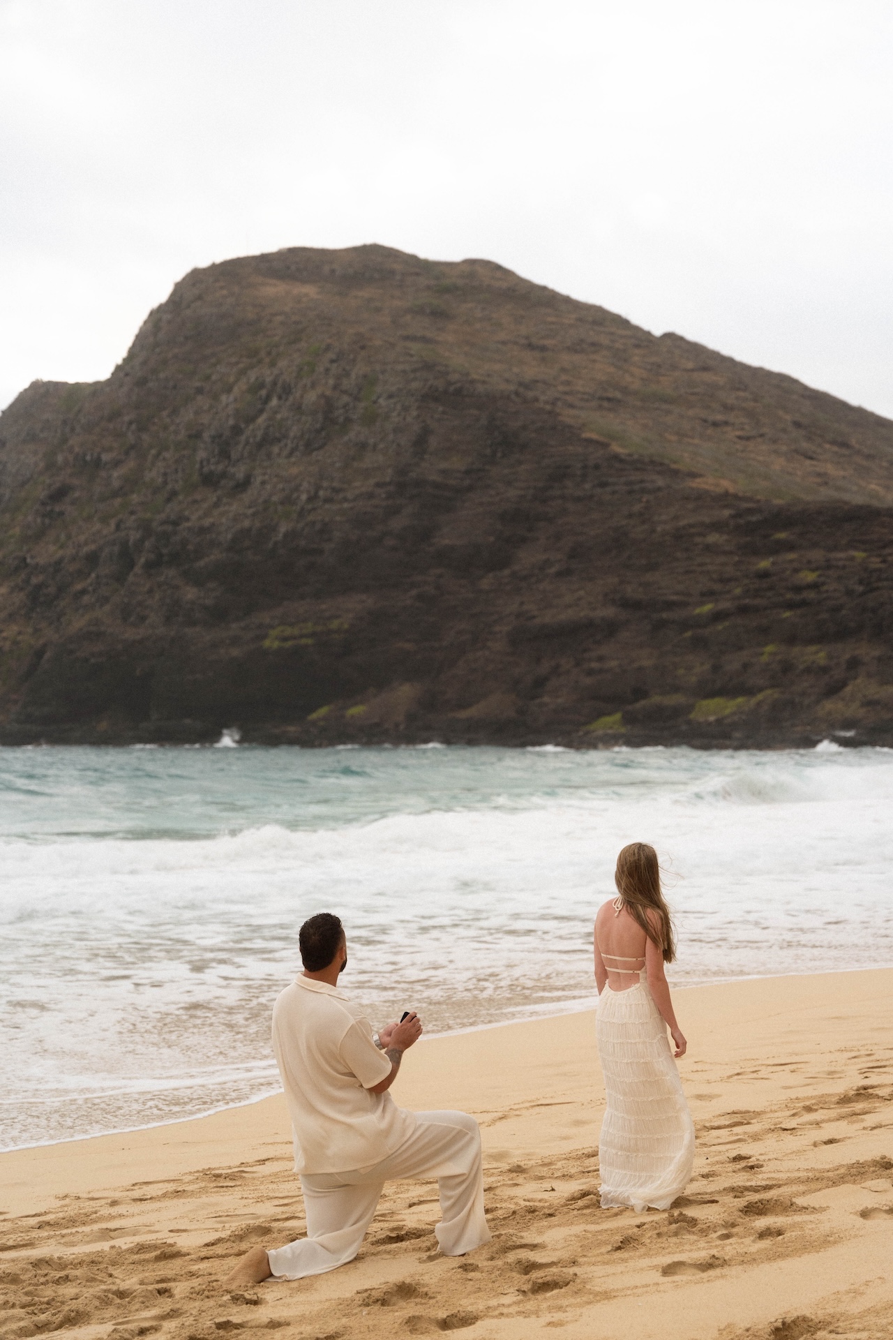 Groom-to-be kneeling on the shoreline holding a ring box, his partner looking out toward the waves.
