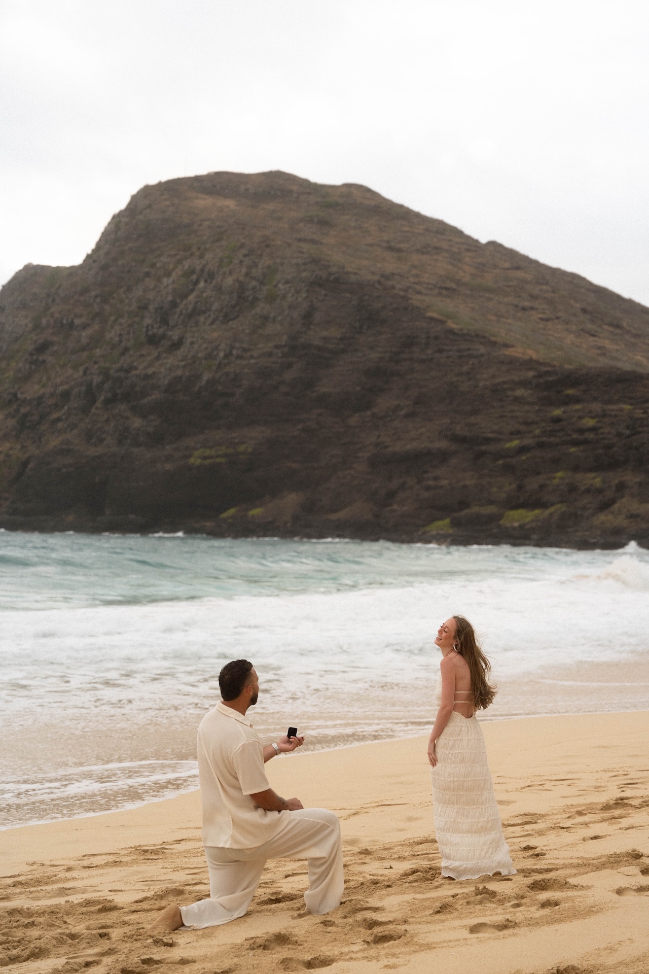 Groom-to-be kneeling on the sand, holding out a ring as his partner smiles joyfully during their beach proposal.