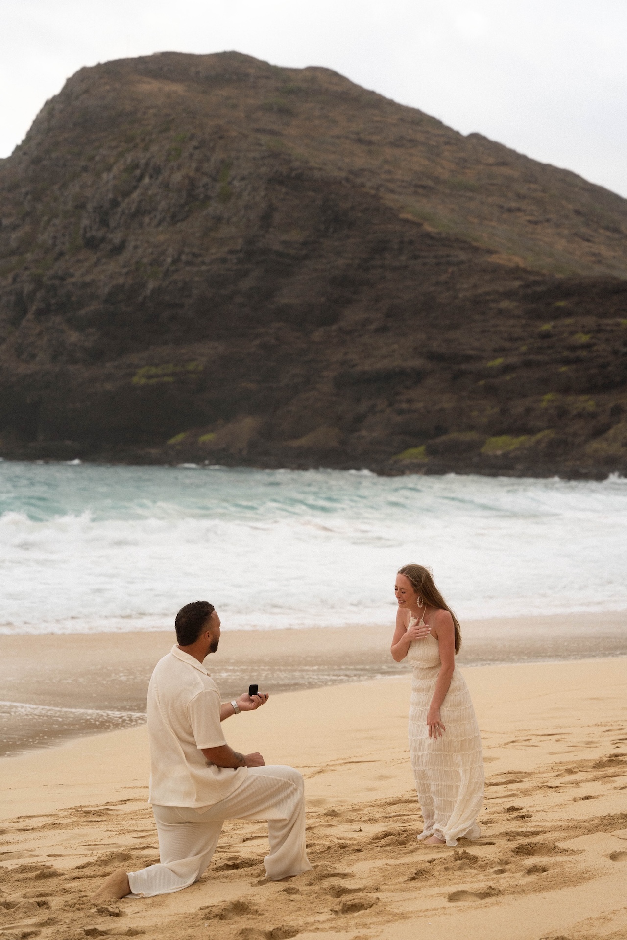 Intimate beach proposal moment as the man kneels with a ring box while his partner reacts emotionally, hand to her chest.