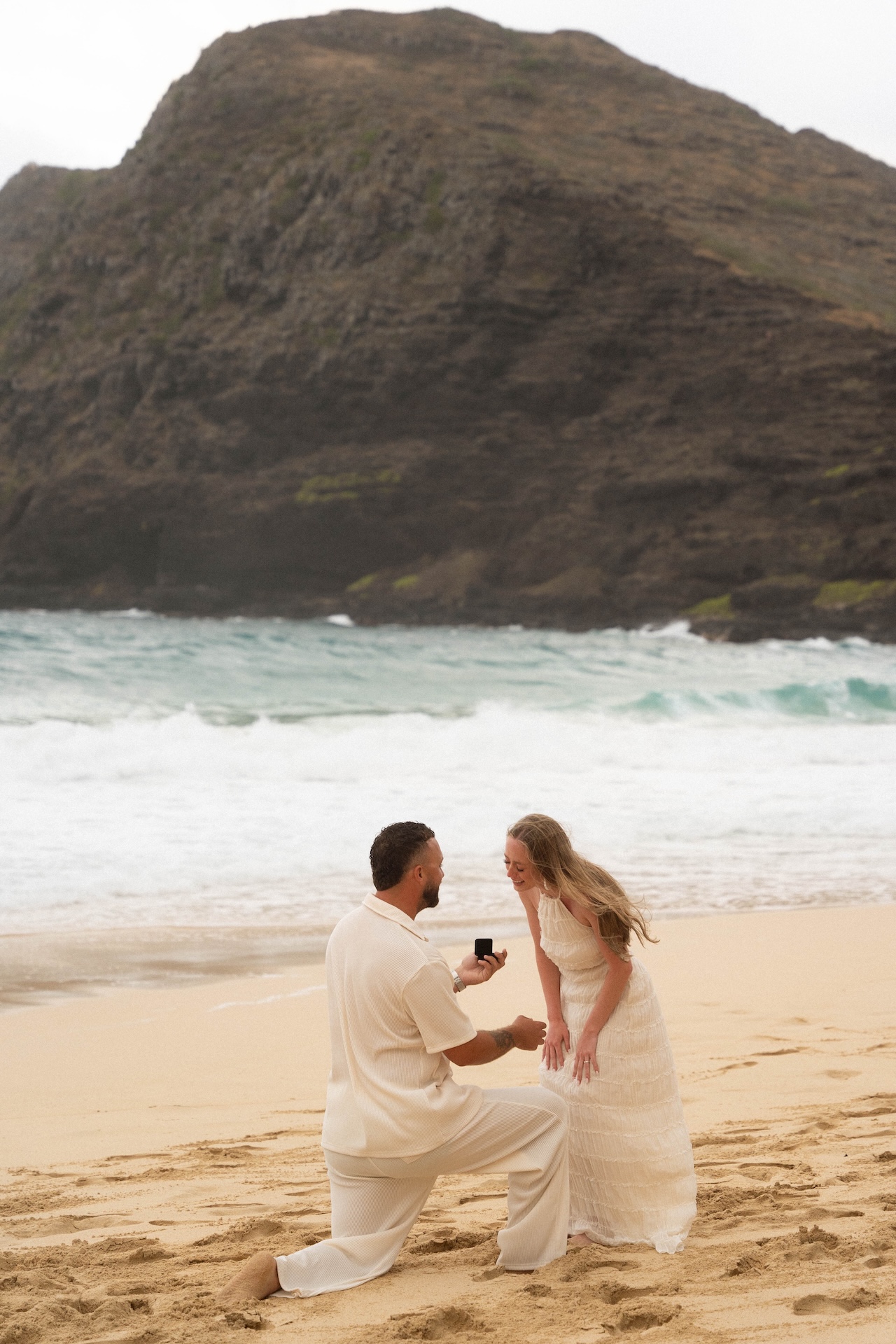 Intimate beach proposal scene where the bride-to-be leans in, smiling and emotional as he presents the ring.