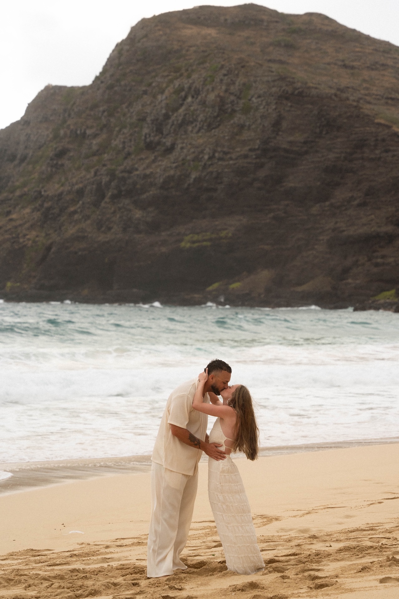 Couple sharing a romantic kiss on the sand with ocean waves and cliffs creating a dramatic backdrop.