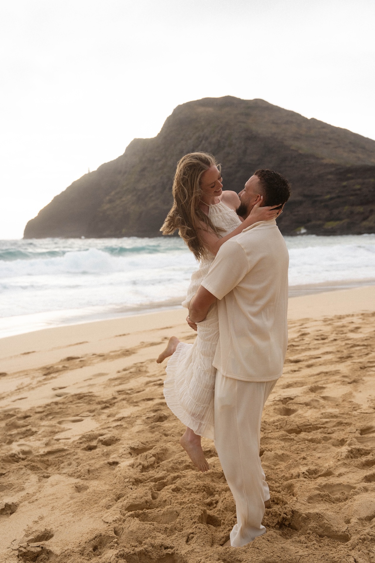 Groom-to-be lifting his fiancée into his arms on the beach, both smiling joyfully as the sun sets on their beach proposal.