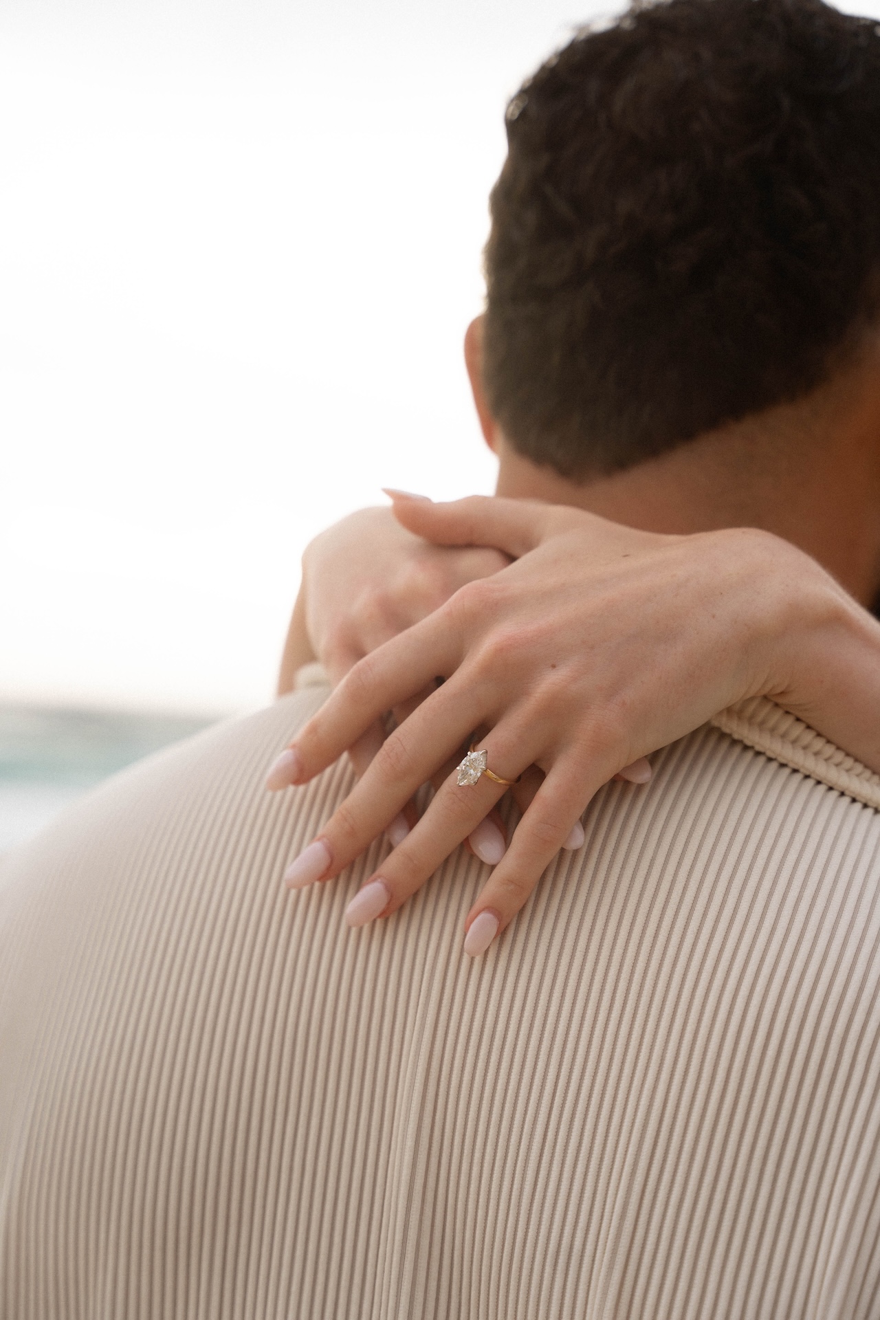 Close-up of the bride-to-be’s hand wrapped around her fiancé’s shoulder, showing the elegant diamond engagement ring from their beach proposal.