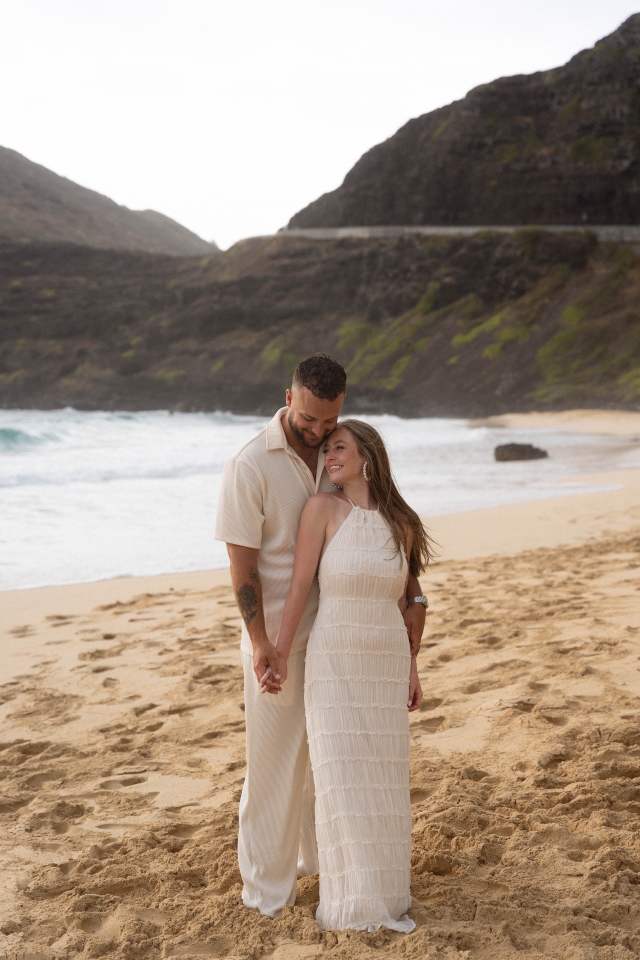 Couple holding hands and smiling at each other on the sandy shoreline, with waves rolling in the background.