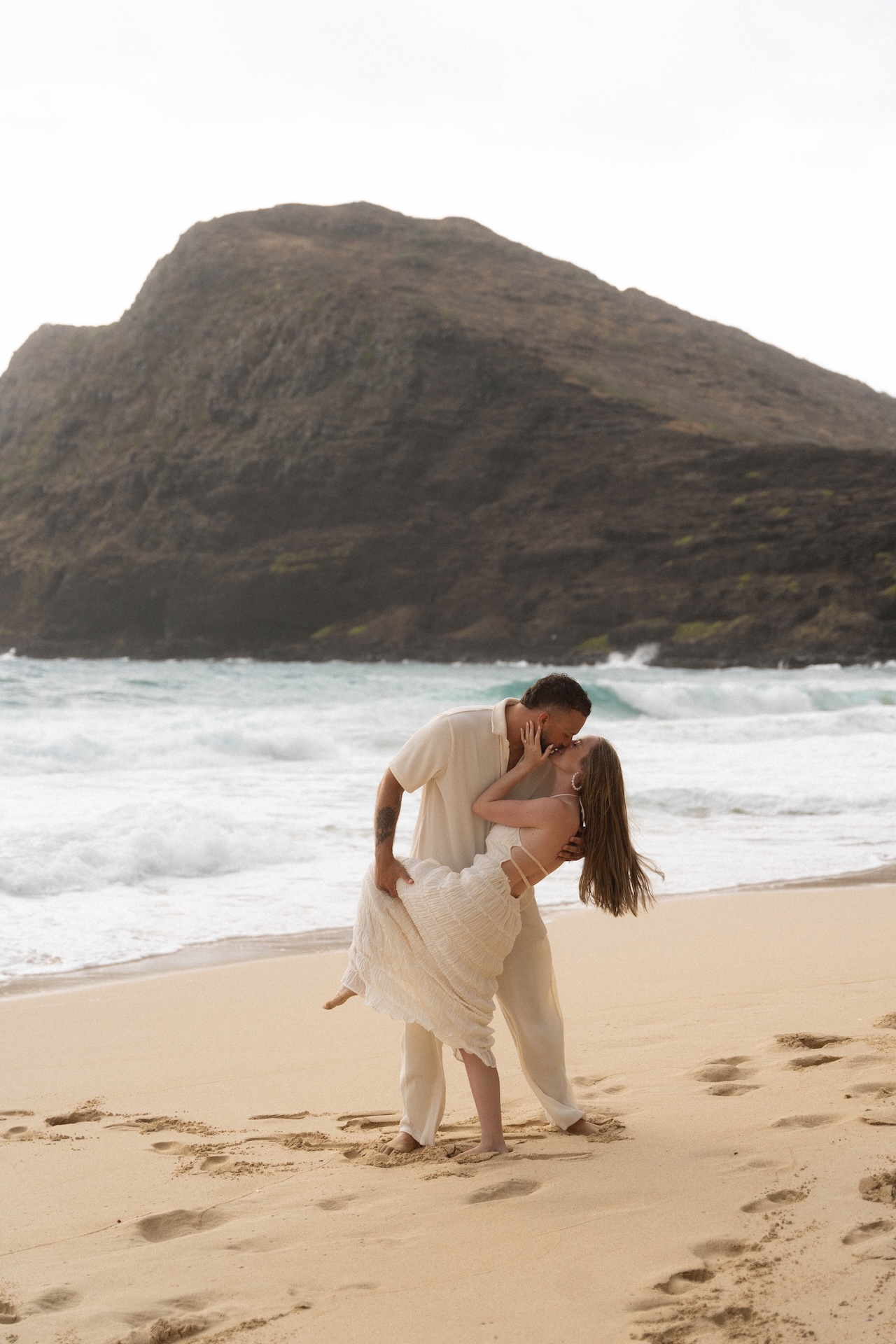 Couple sharing a passionate dip-and-kiss on the beach, framed by ocean waves and rugged cliffs in the background.