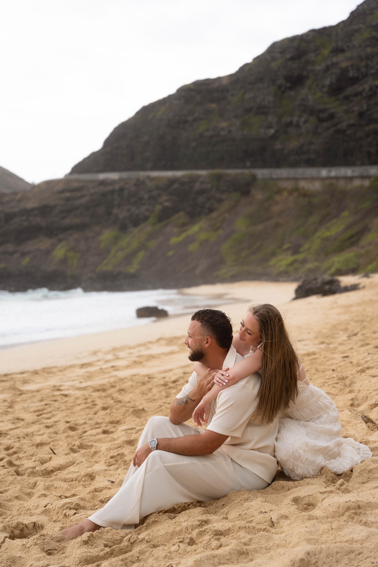 Bride-to-be sitting behind her fiancé on the sand, wrapping her arms around him in a sweet and intimate moment during their beach proposal.