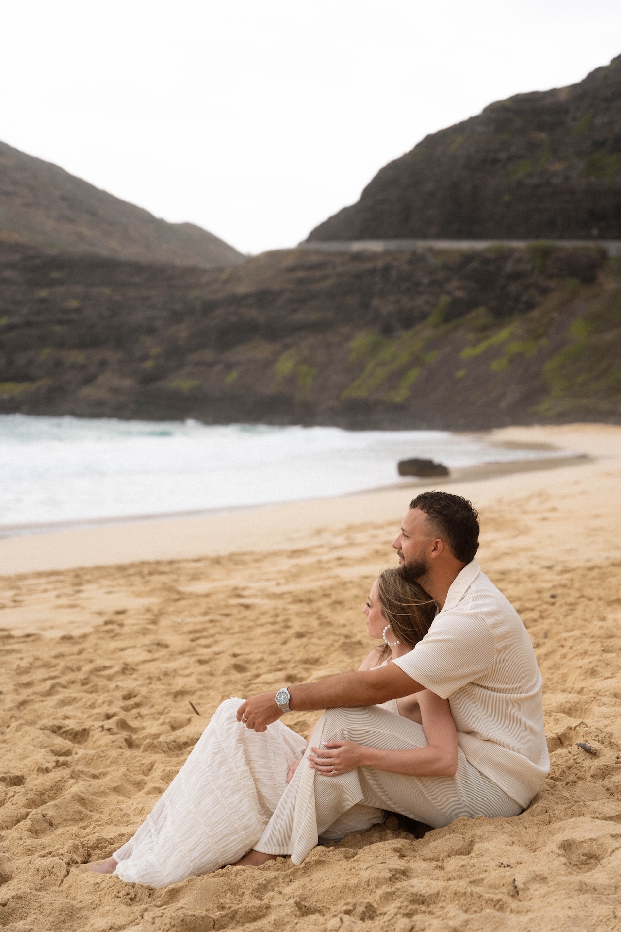 Romantic scene of the couple sitting close together on the sand, leaning into each other while gazing out at the ocean waves.