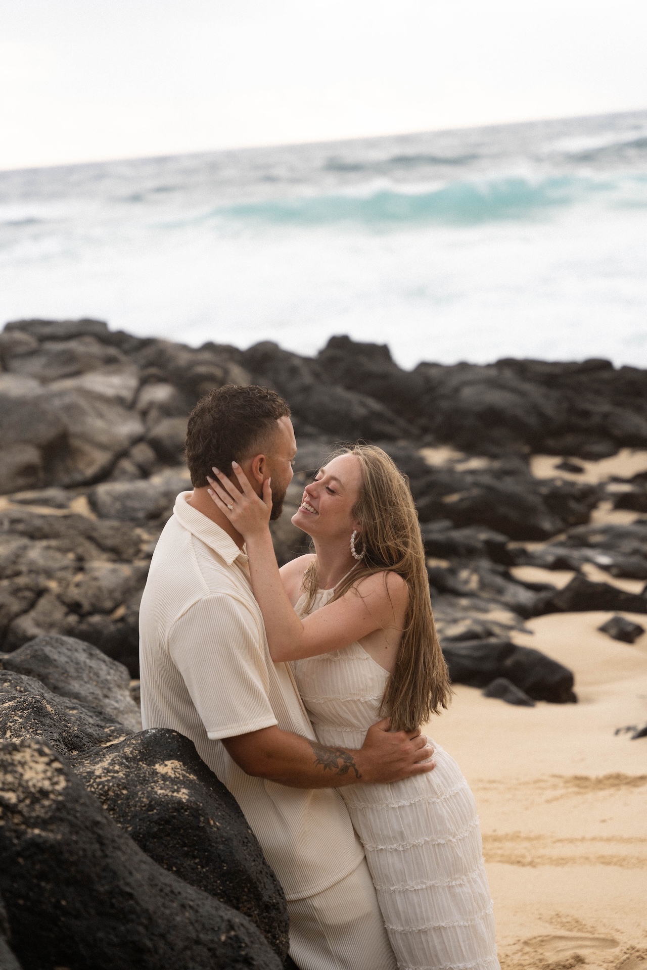 Romantic portrait of the couple embracing by the lava rocks, smiling lovingly at each other during their beach proposal session.
