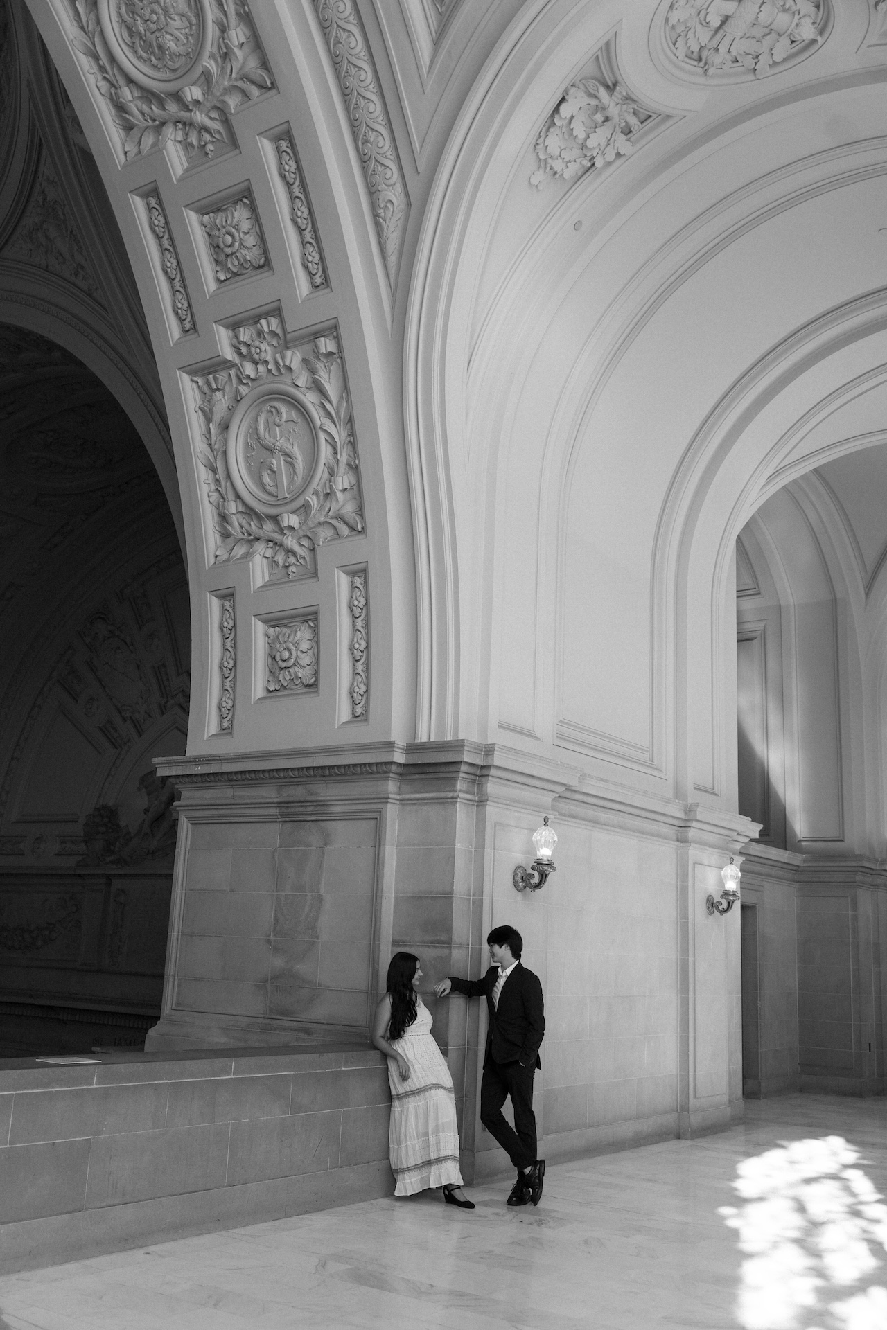 A stunning vertical photo of the bride and groom smiling at each other and leaning against the stone railing at the San Francisco City Hall with a view of the rotunda above them.