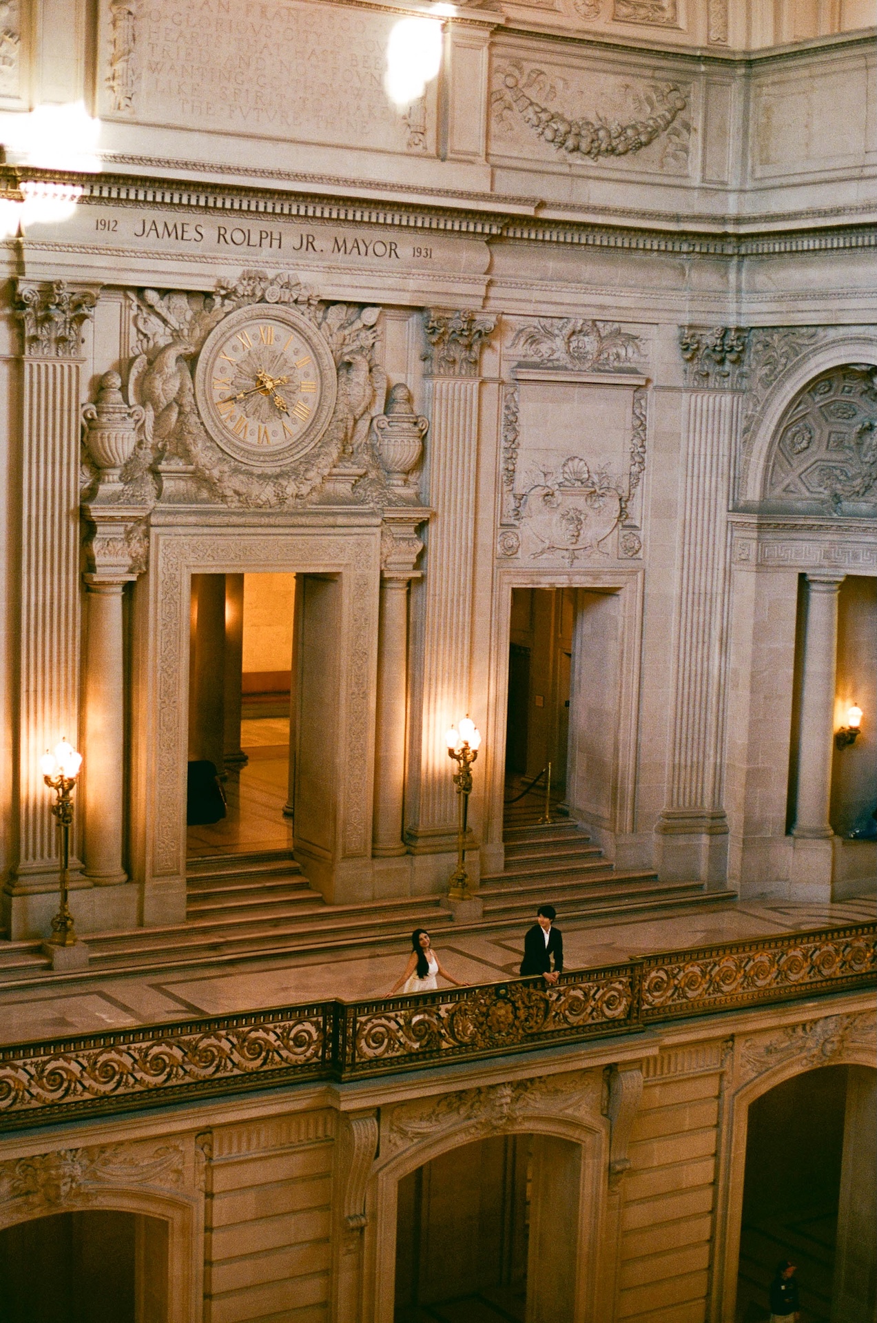An overhead shot shows a couple standing on a balcony near a large decorative clock, a classic backdrop for a San Francisco City Hall wedding.