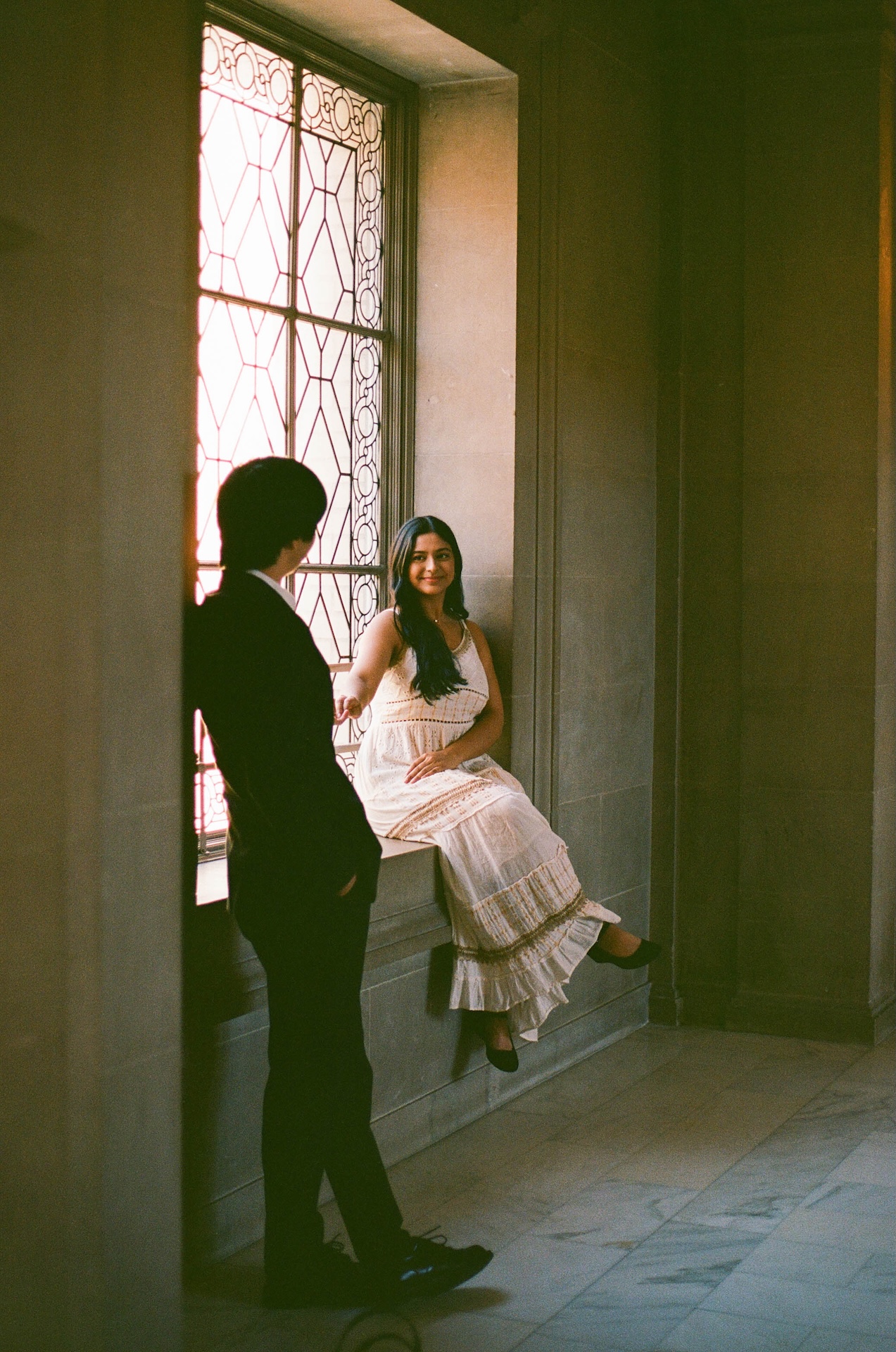 A woman sits on a sunlit window ledge while the man stands next to her, as if in mid-conversation.