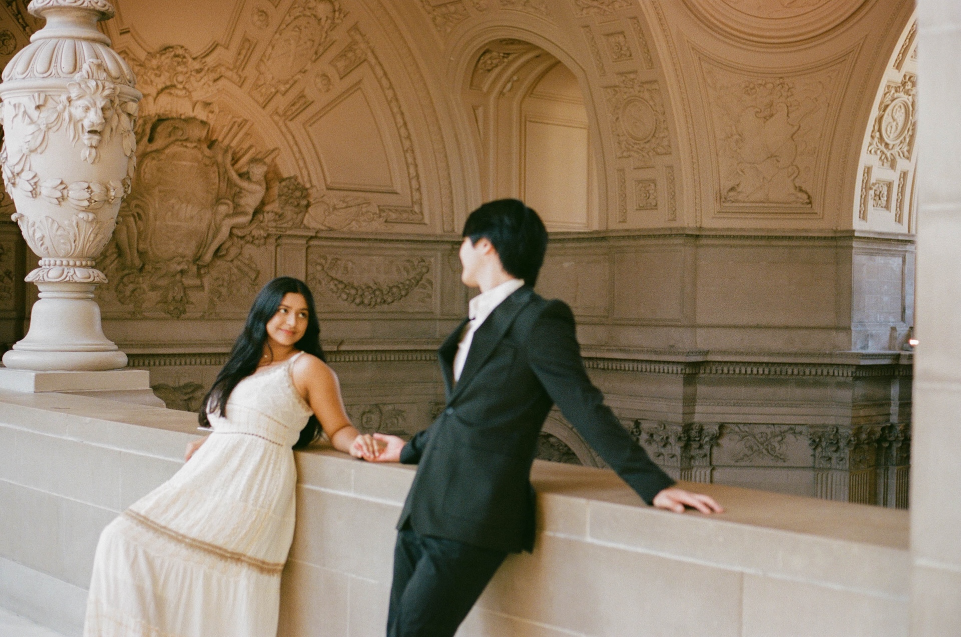 A candid shot of a couple leaning against a stone railing in a magnificent, ornately decorated hall. They are holding hands and smiling at each other.