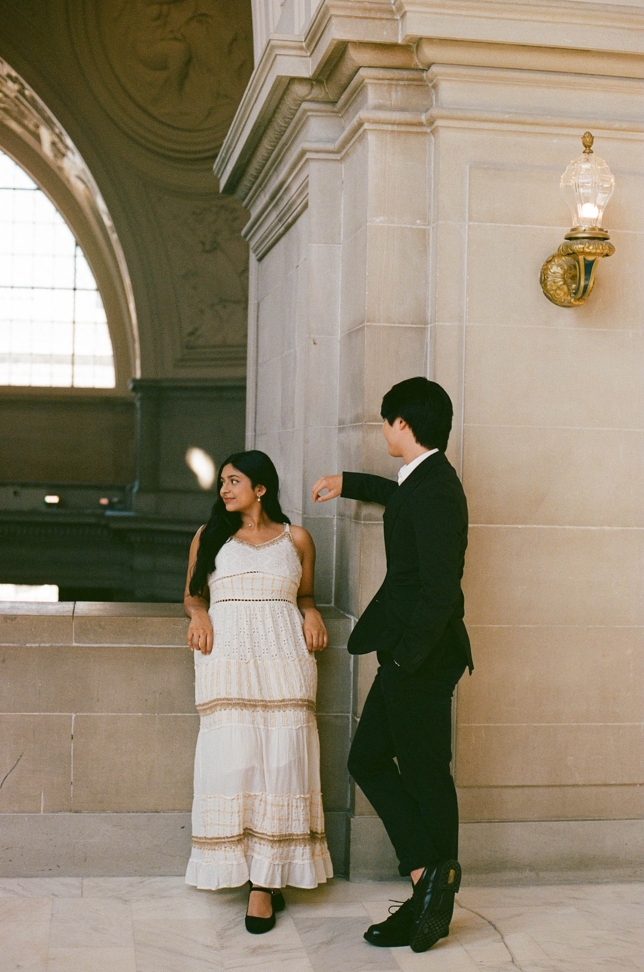 A couple stands together in a hallway, leaning against a pillar and looking at each other.