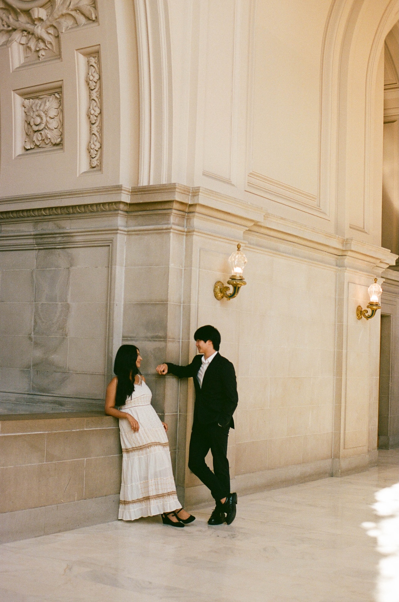 A couple poses while sitting on a balcony ledge, with the elaborate arches and carvings of the building in the background. A beautiful shot from their San Francisco City Hall wedding.