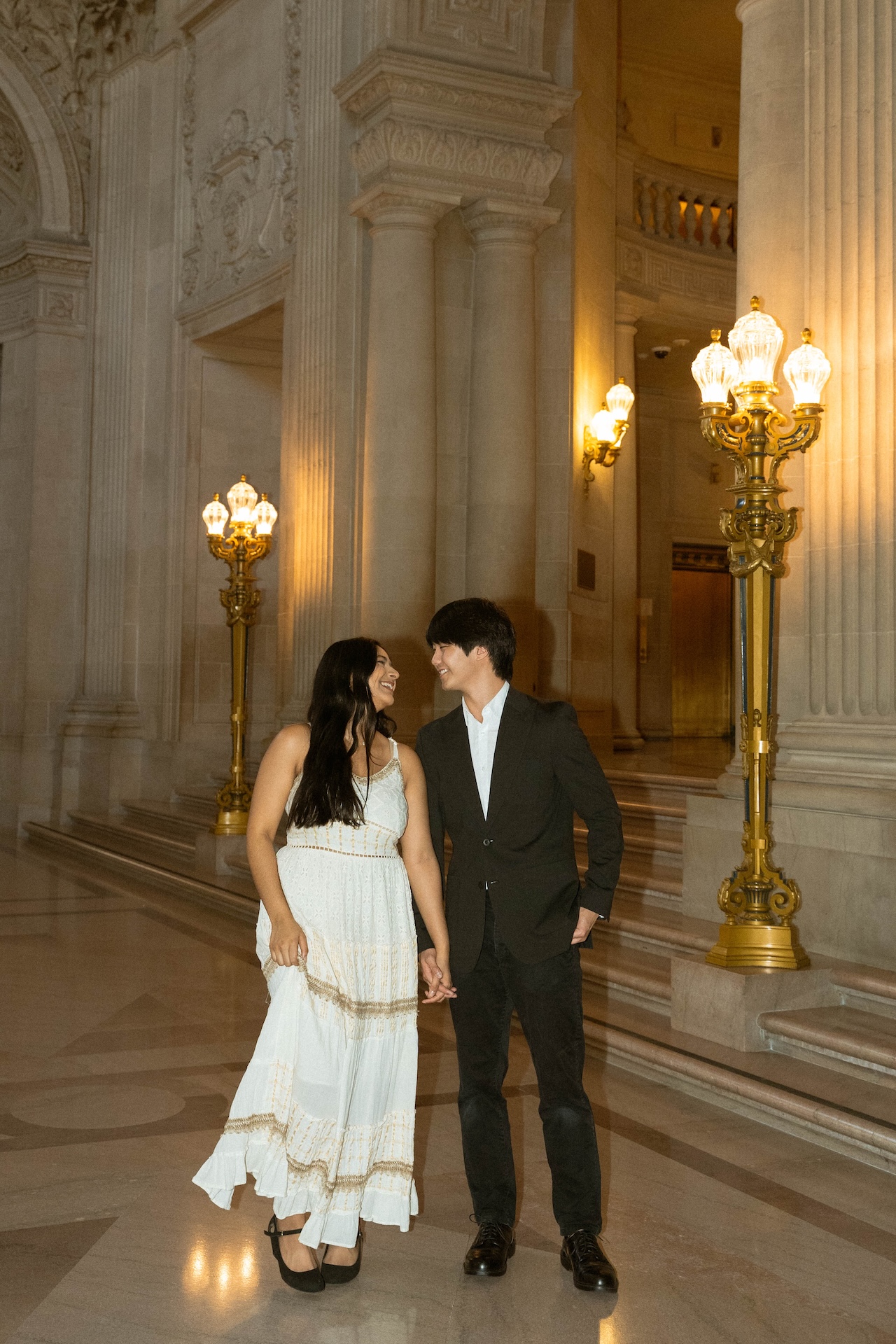 A couple stands holding hands in a brightly lit hall with tall columns and intricate light fixtures. They are smiling and looking at each other.