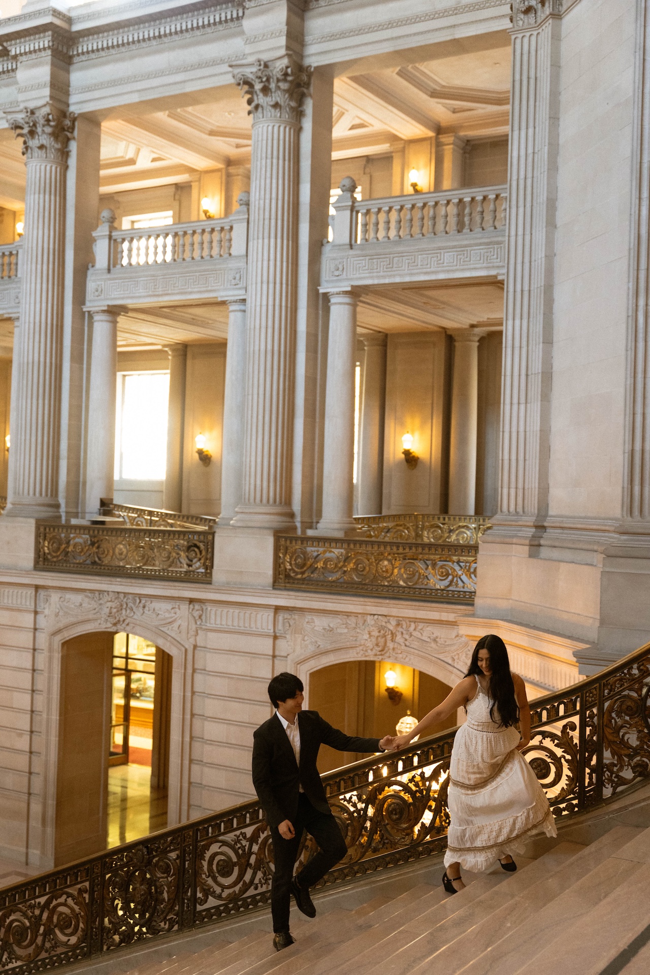 A couple walks up a grand staircase in a massive, ornate building, holding hands and looking at each other.