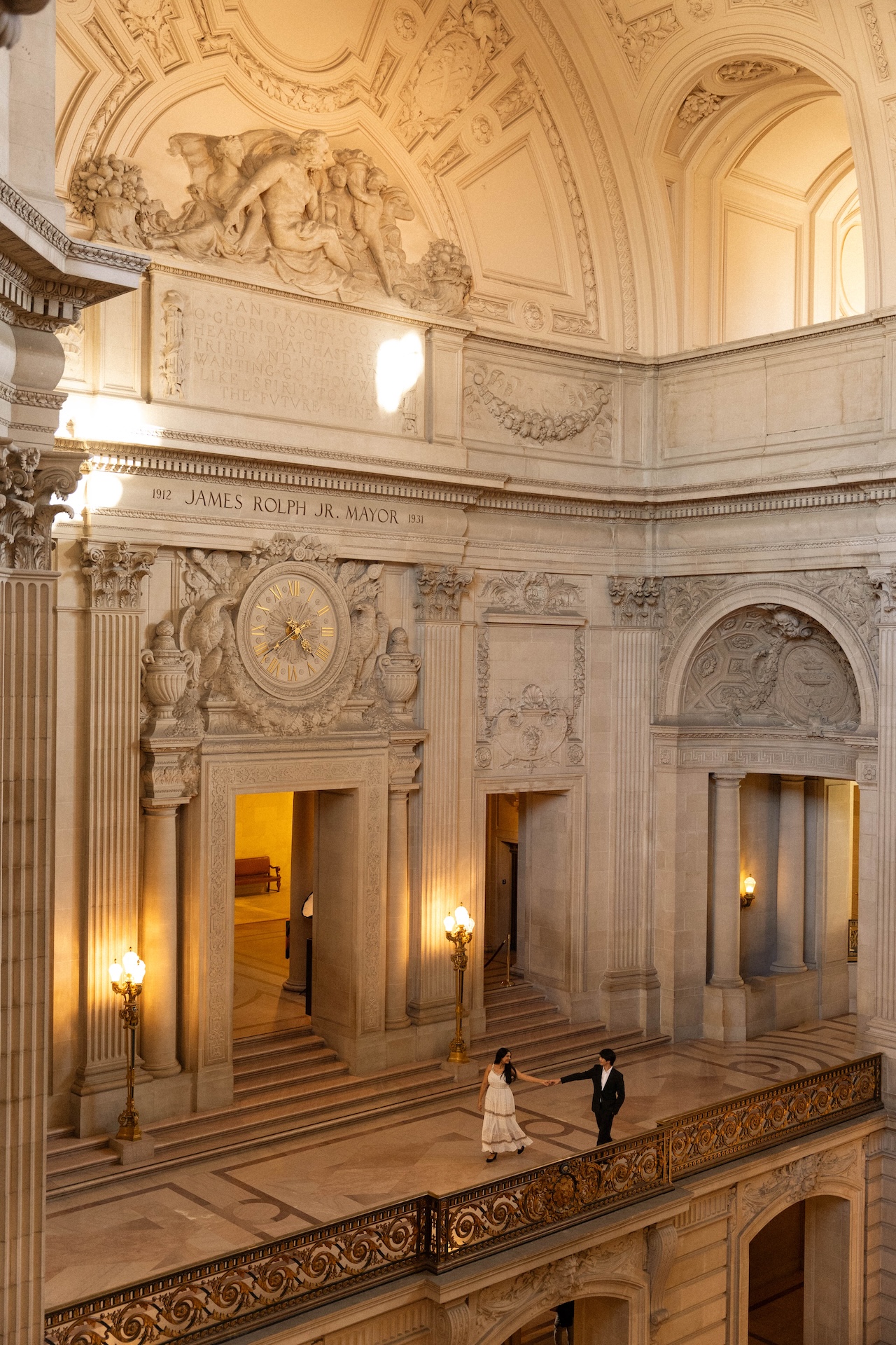 A couple walks down a grand staircase in a massive, ornate building, holding hands and looking at each other.