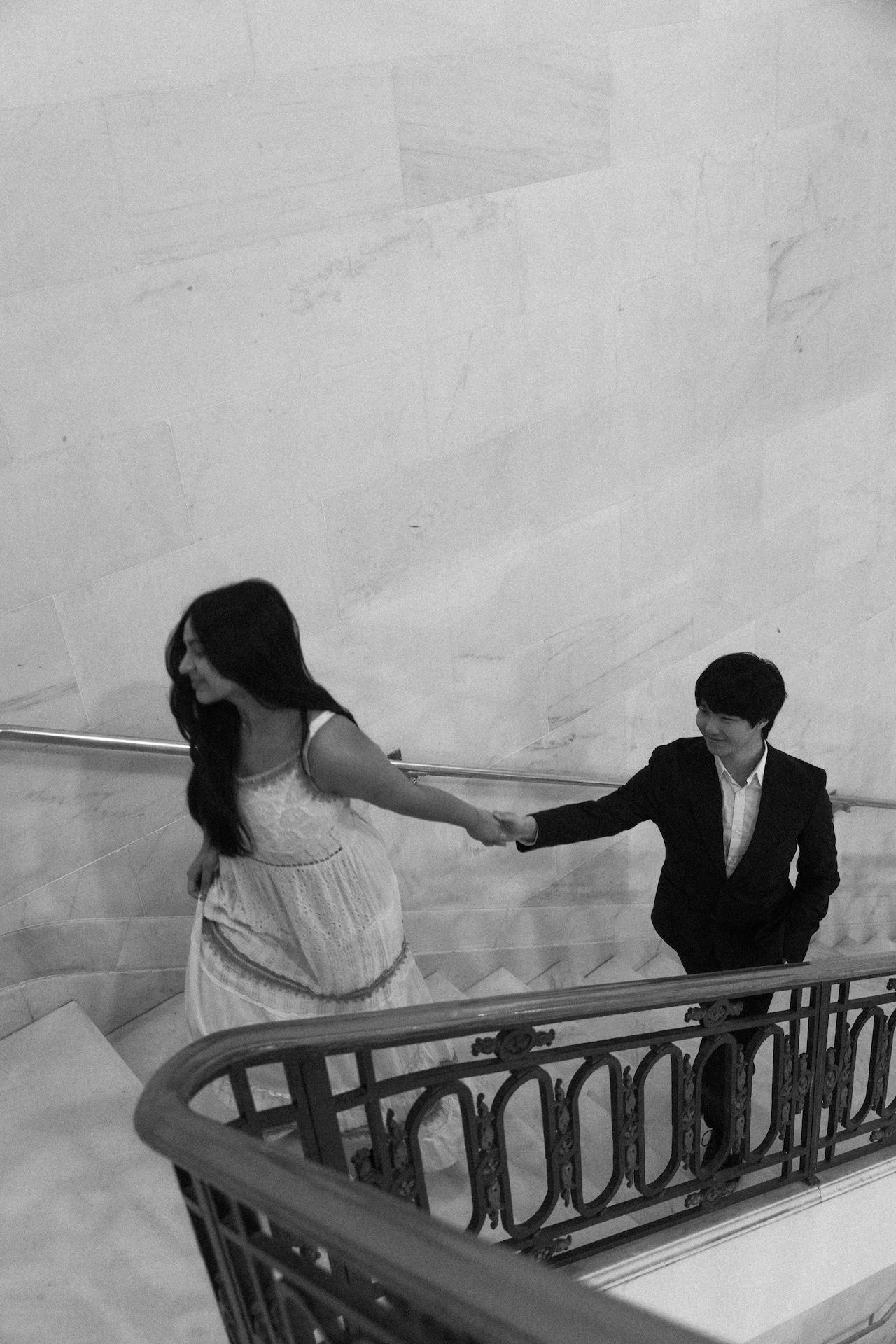 A black and white photo of a couple walking up a staircase, holding hands and looking back. This is a romantic moment from a San Francisco City Hall wedding.