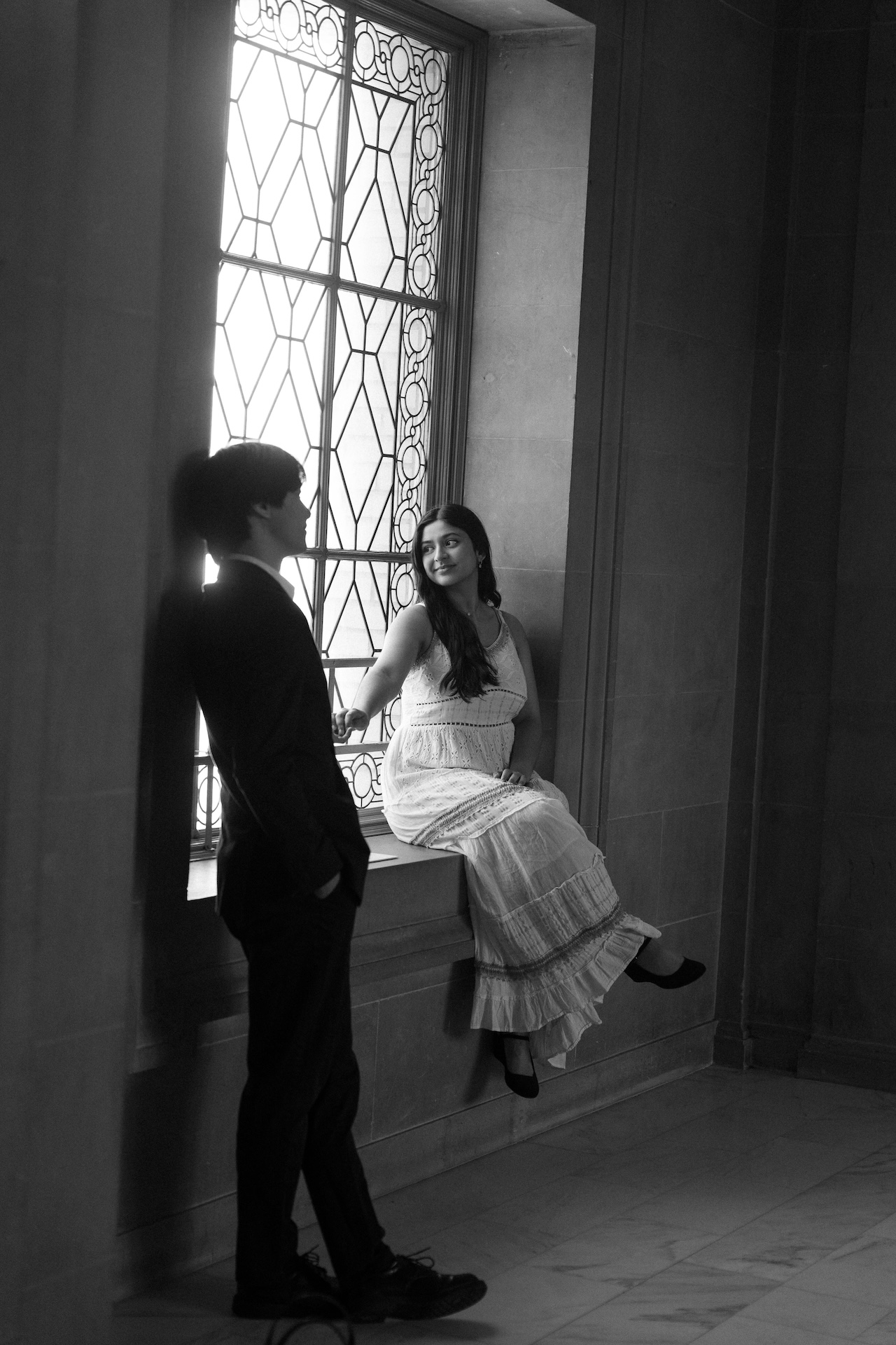 A black and white image of a bride sitting on a sunlit window ledge while the groom stands next to her.