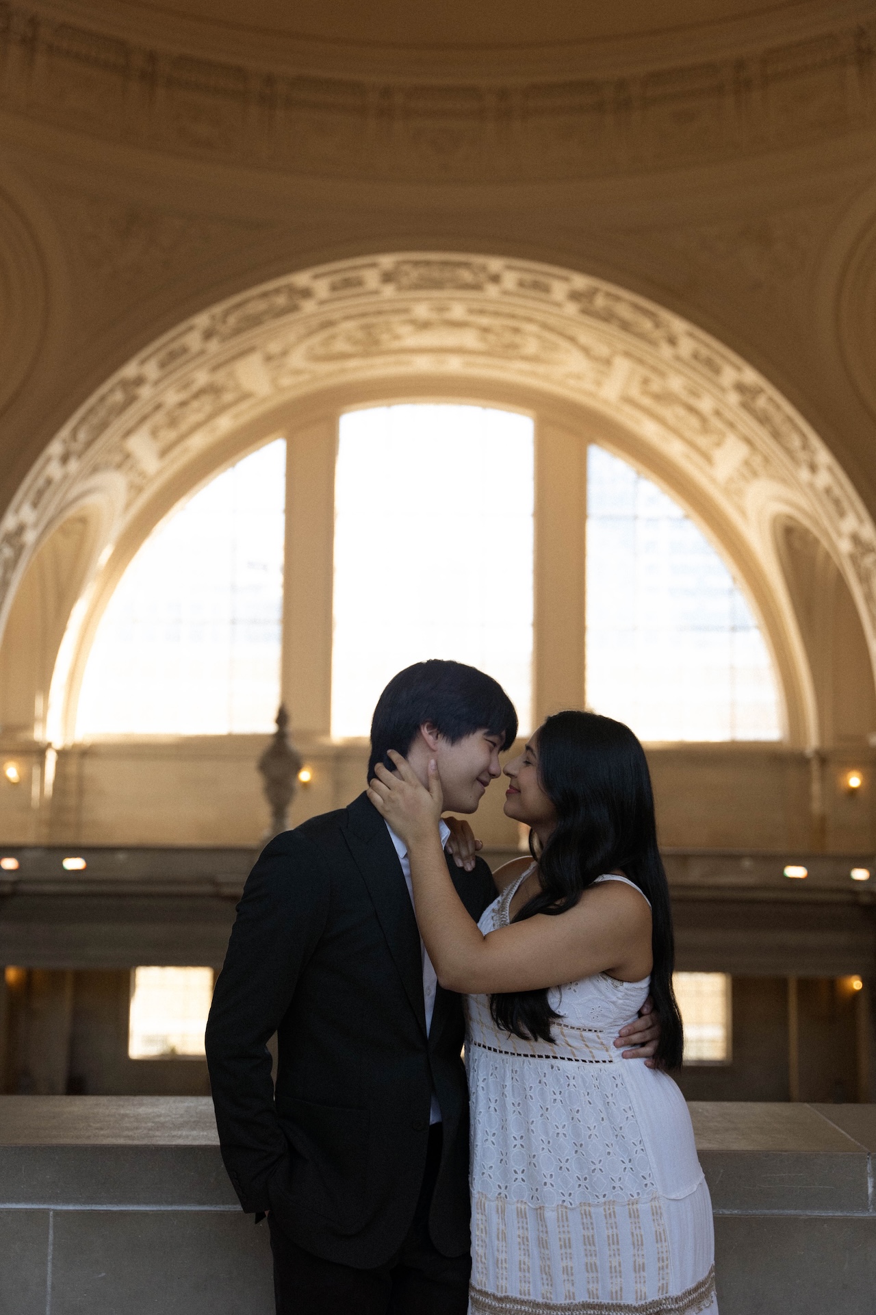A romantic close-up of a couple, facing each other and smiling. They are leaning against a ledge with large, sunlit windows in the background.