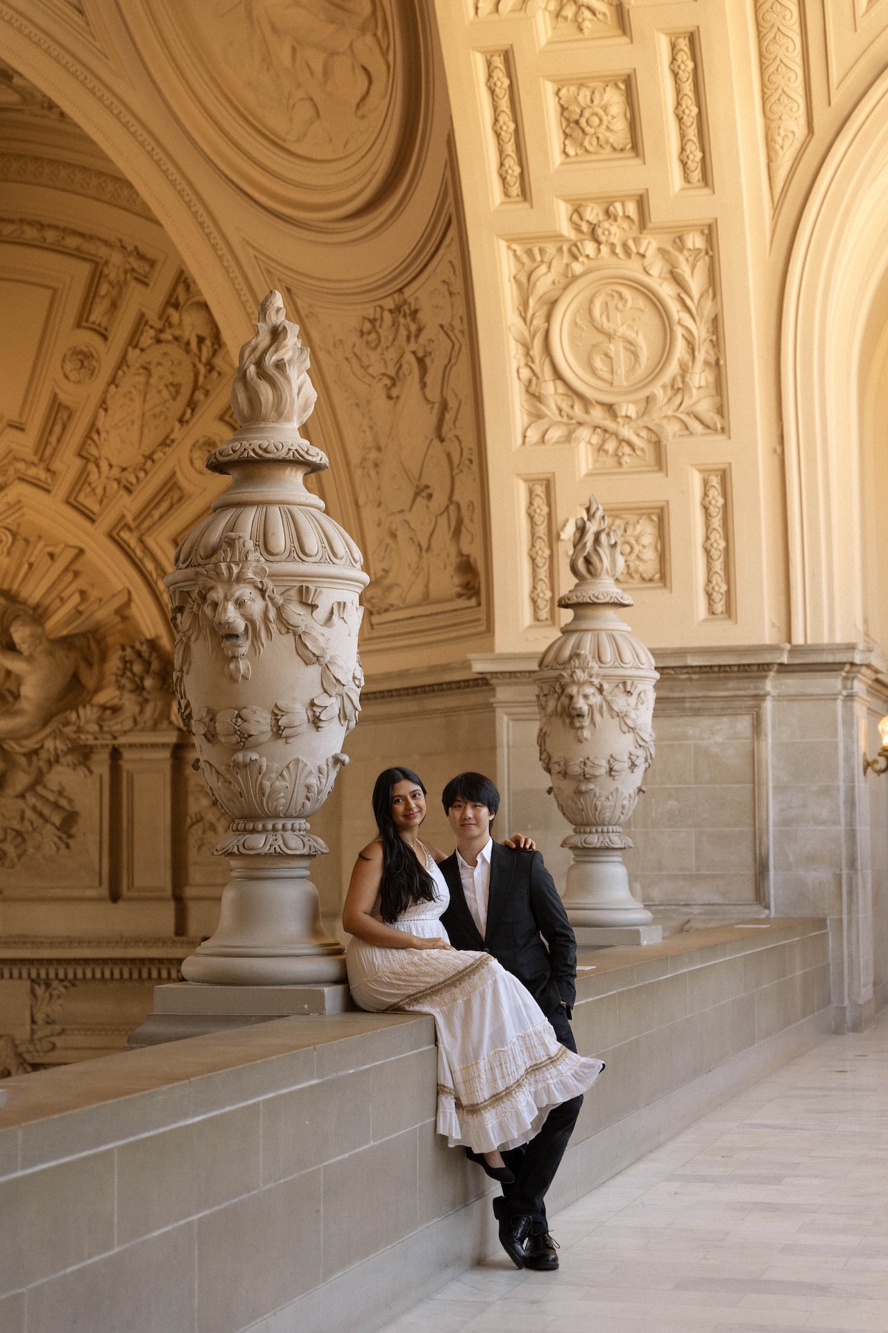 The bride seating on the ledge of the stone railing while the groom holds her. Both smiling softly at the San Francisco City Hall