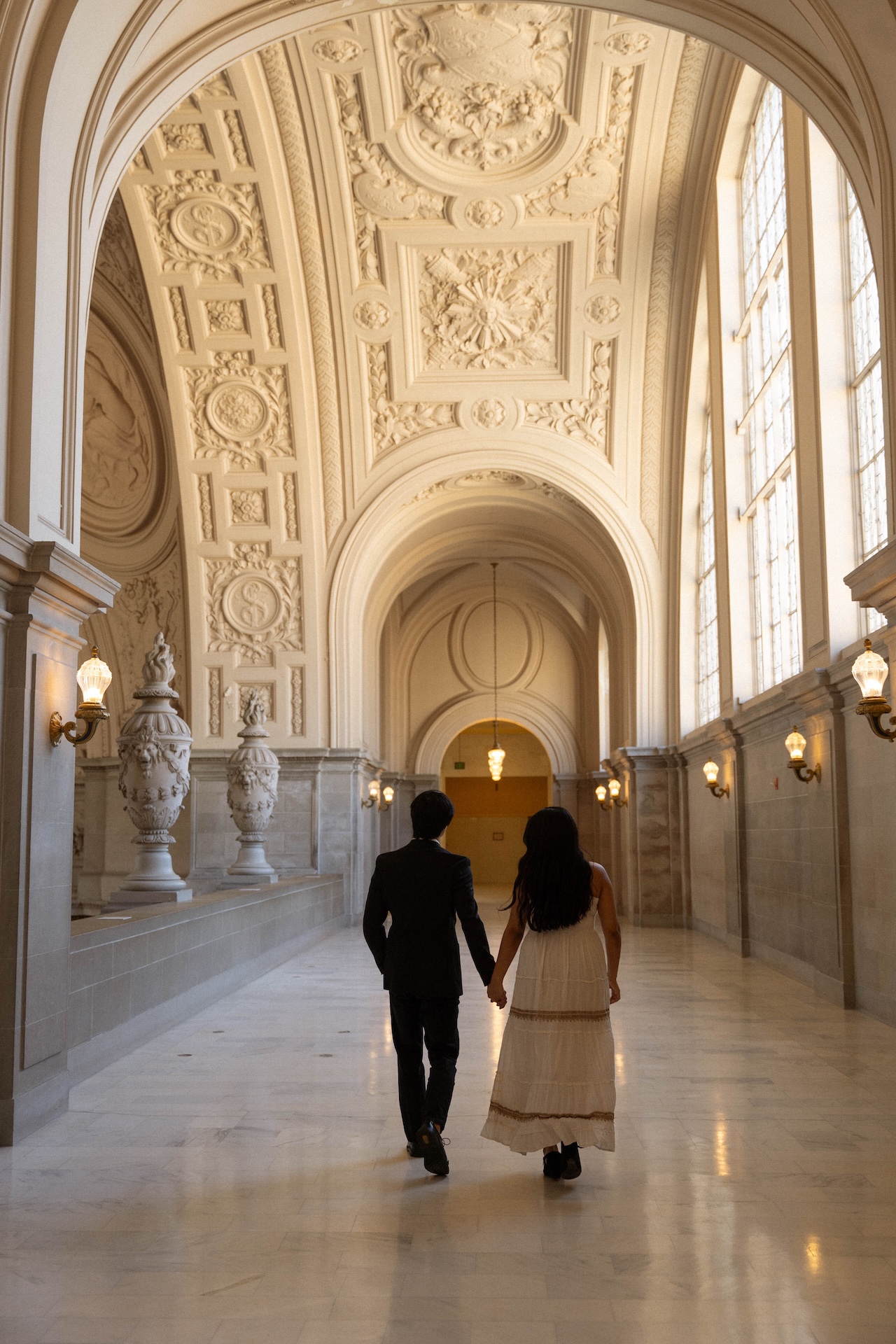 A couple holding hands and walking away down a long, elegant hallway with a decorative, vaulted ceiling.