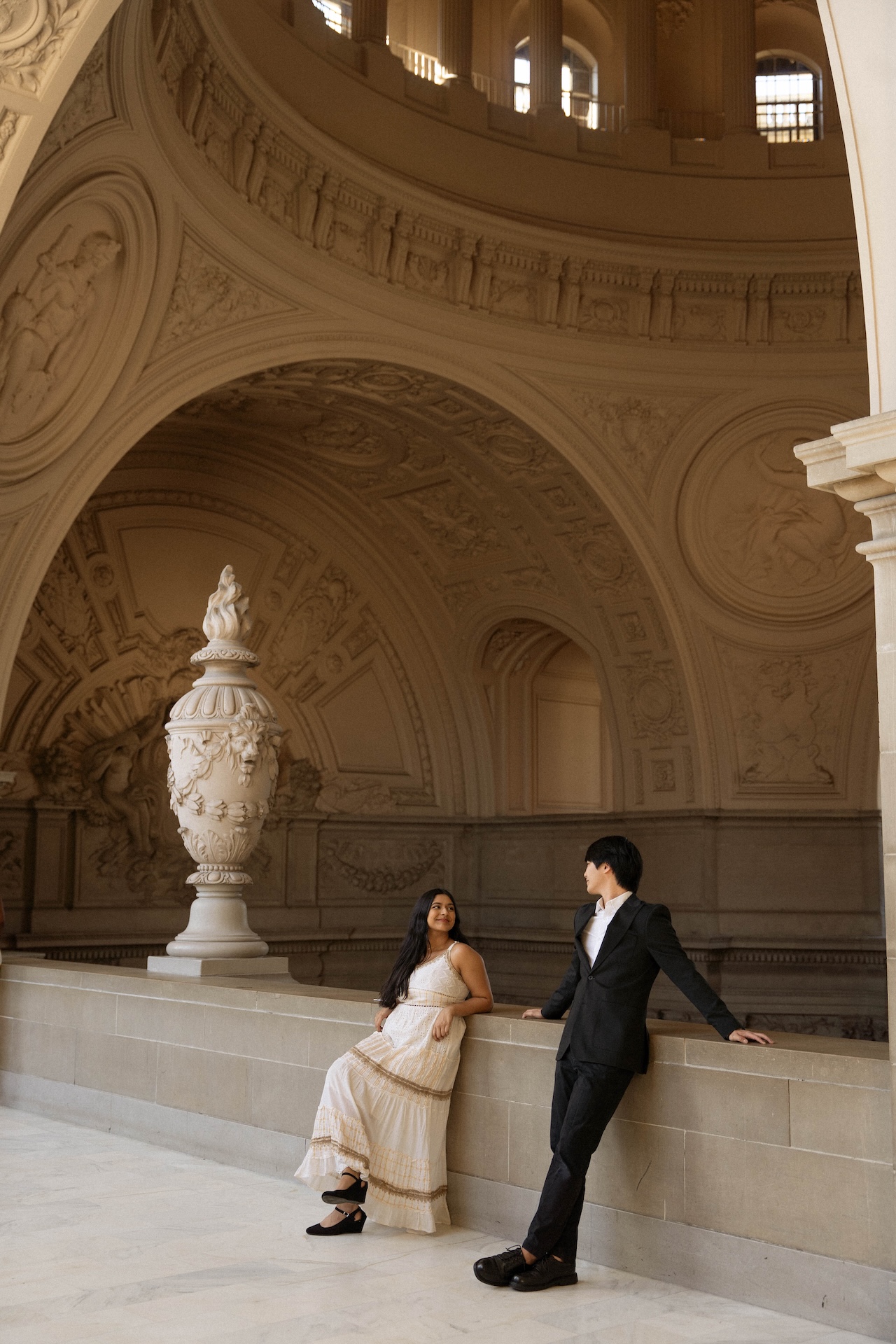 A stunning vertical photo of the bride and groom smiling at each other and leaning against the stone railing at the San Francisco City Hall with a view of the rotunda above them.