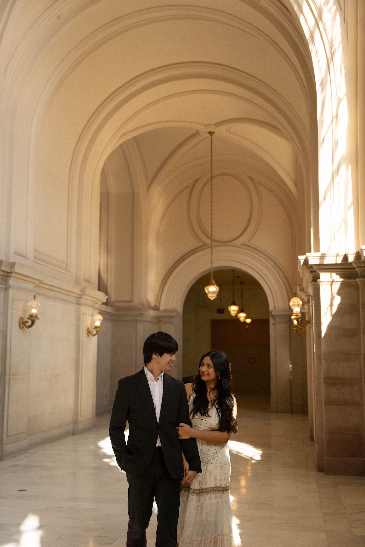 The bride and groom stand on a balcony overlooking the grand interior of a public building. They are looking down and holding hands.