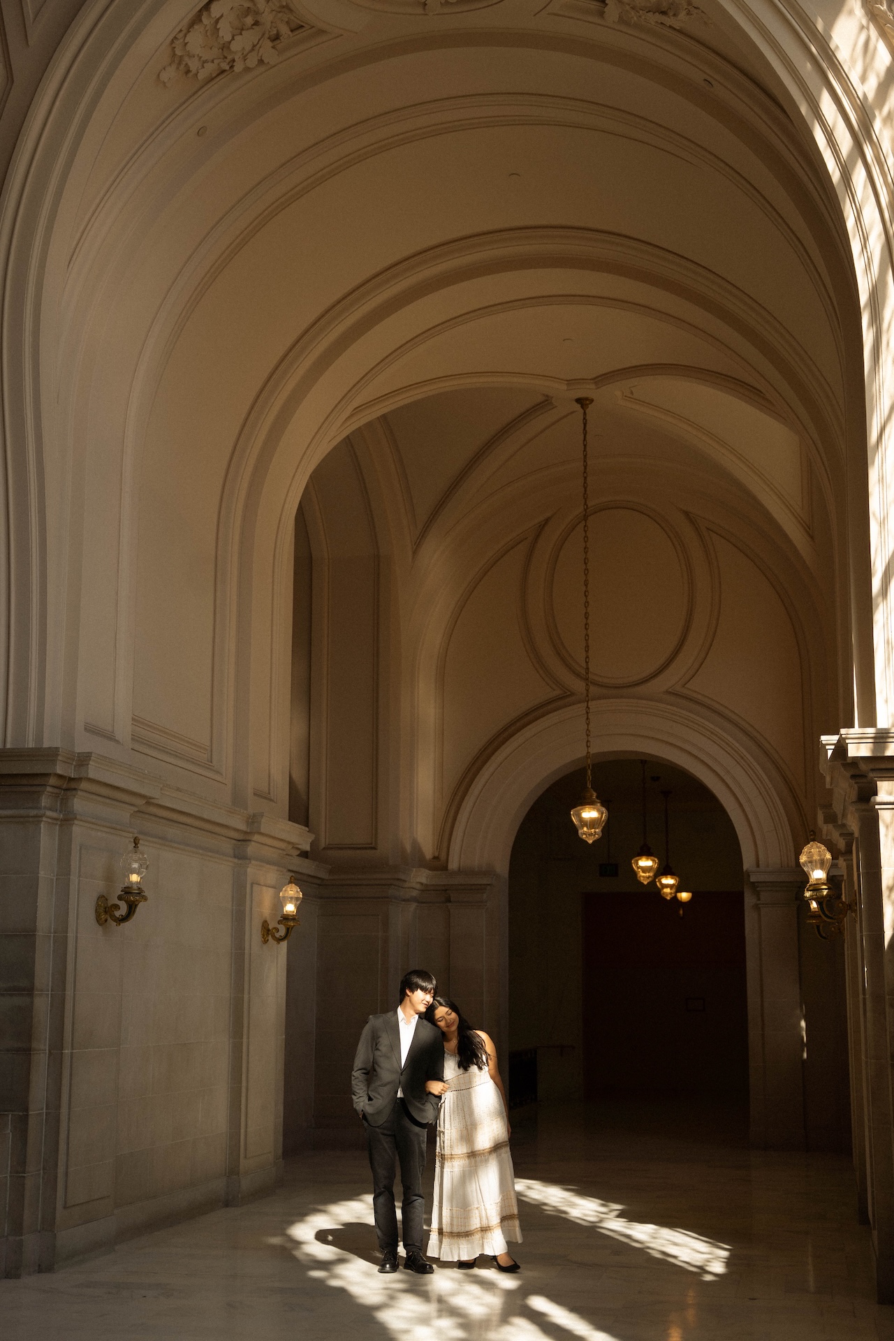 A couple stands together in a sun-drenched hallway, their bodies close together in a sweet embrace.