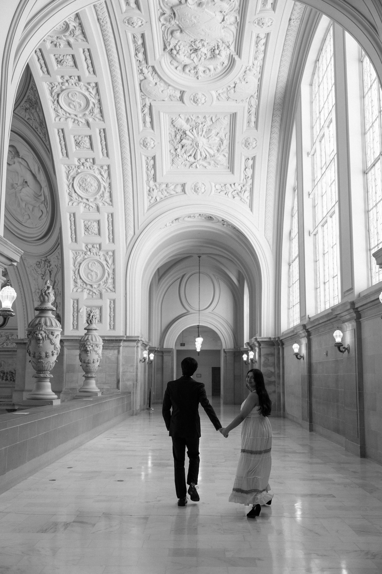 A black and white photo of a couple holding hands and walking away down a long, elegant hallway with a decorative, vaulted ceiling.
