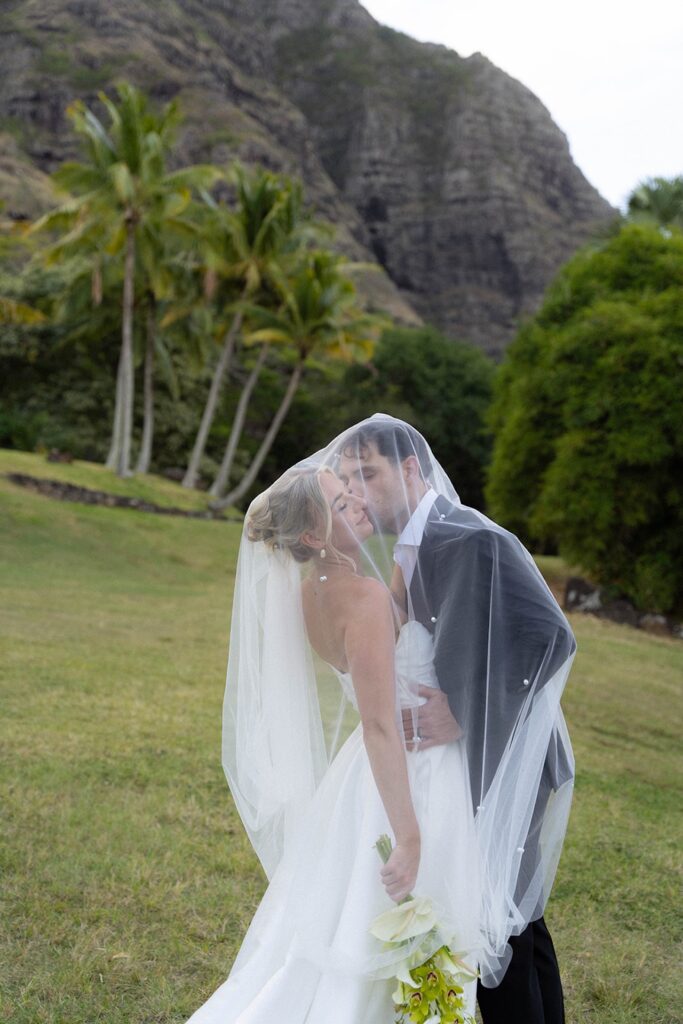 Bride and groom share a romantic kiss under a wedding veil with lush Hawaiian mountains behind them