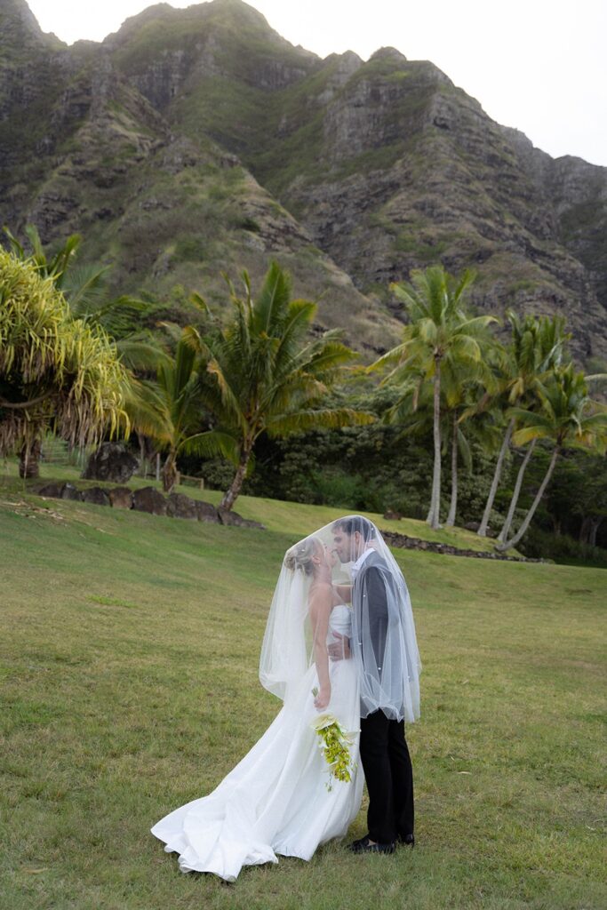 Bride and groom share a quiet kiss under the veil with towering mountains and swaying palms in the background.