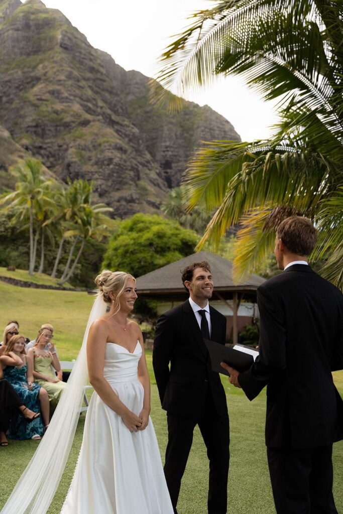 Couple stands before the officiant smiling during their wedding ceremony under palm trees and mountain backdrop