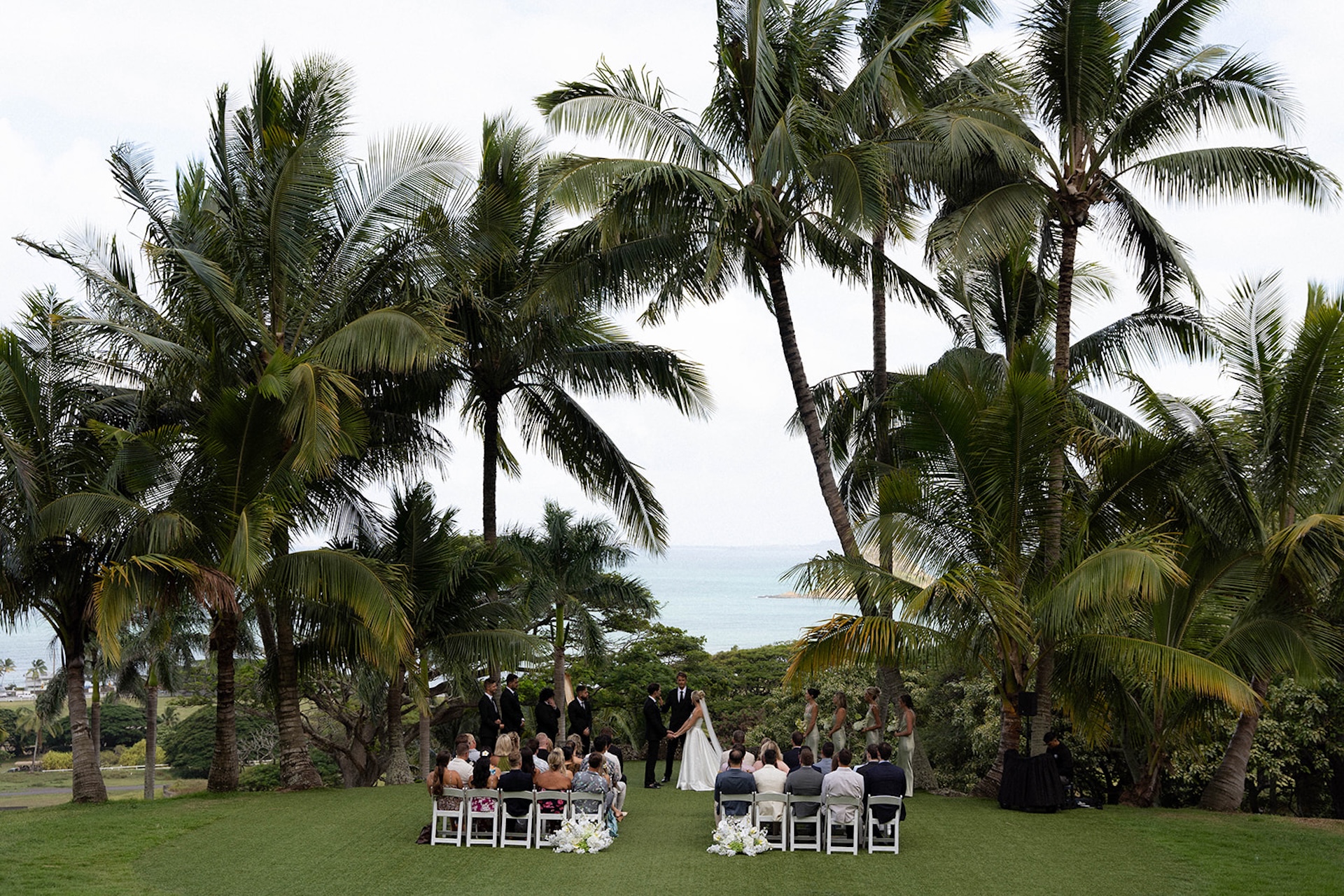 The wedding ceremony unfolds under tall palm trees, with guests seated facing a turquoise ocean backdrop.