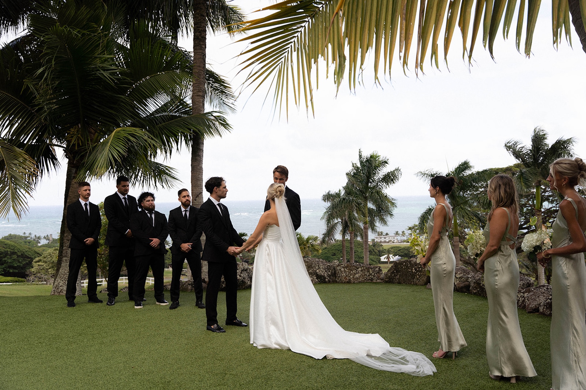 Bride and groom exchange vows in front of their wedding party with panoramic ocean views and tropical palms behind them.
