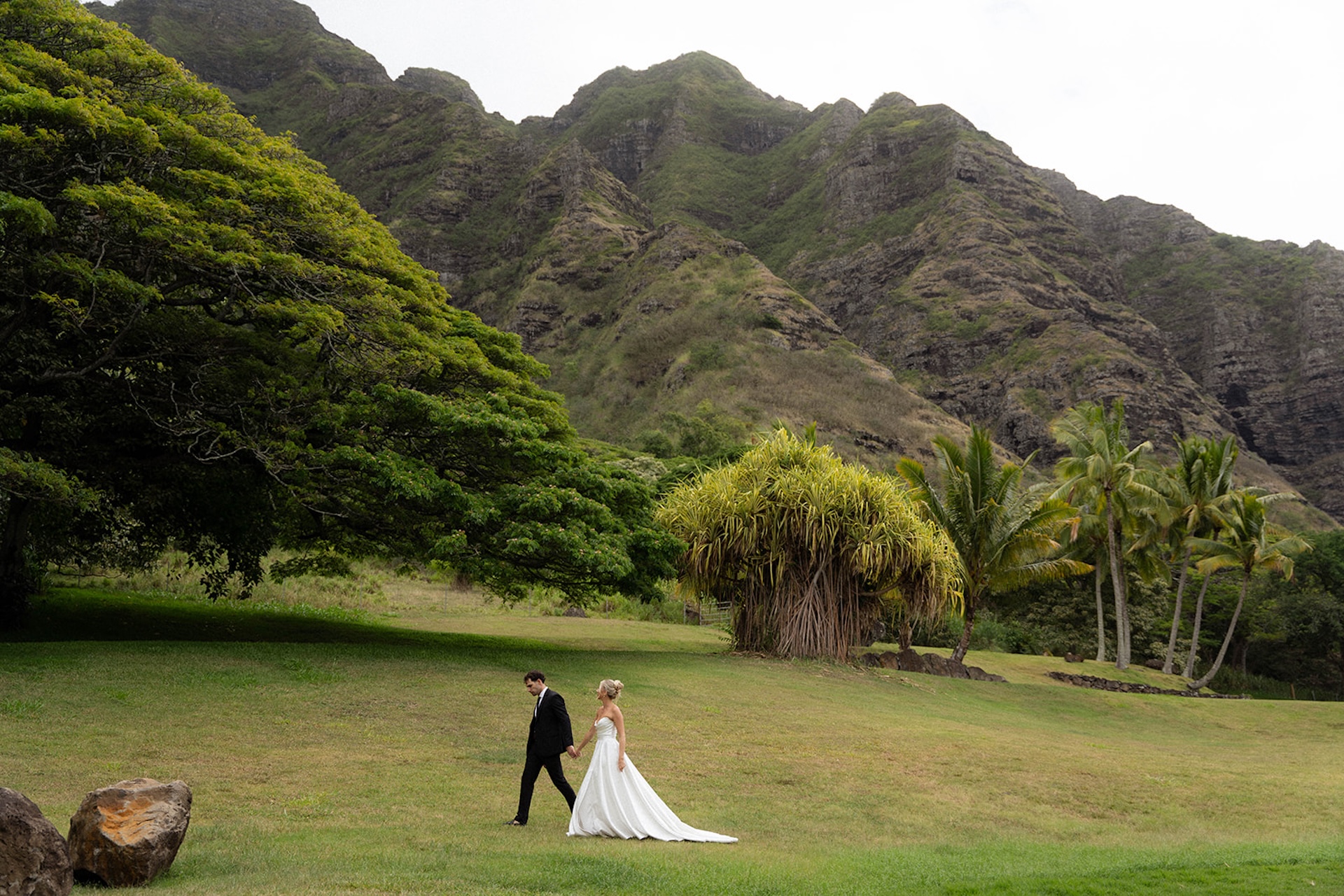 Bride and groom walk across an open field toward towering green mountains – serene moment in Hawaii wedding planning.