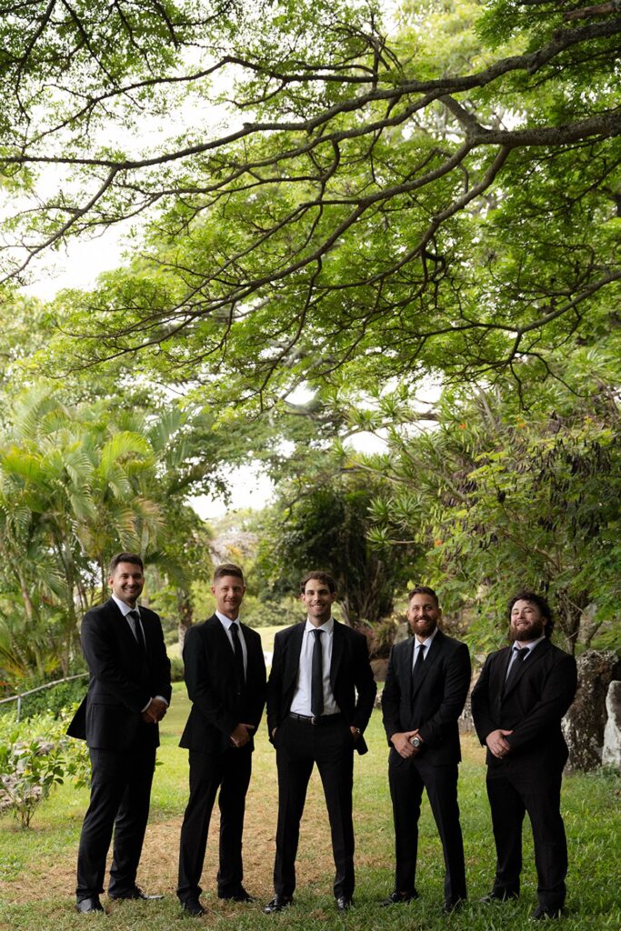 Groom and groomsmen pose together in classic black suits under lush tropical trees before the ceremony.