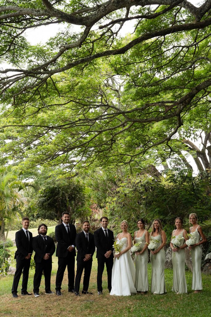 Bridal party poses in formal wear under a massive canopy tree in a lush garden setting.