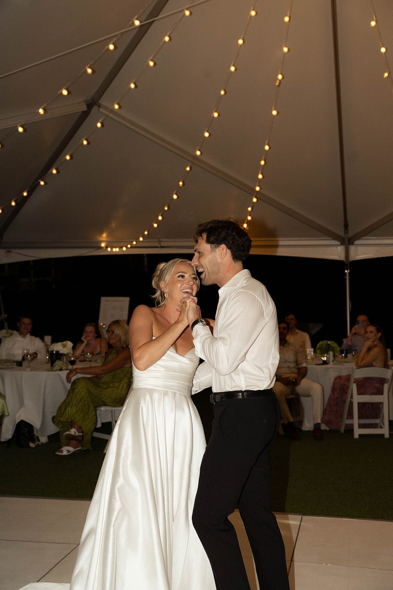 Couple shares their first dance beneath glowing string lights in an elegant reception tent.
