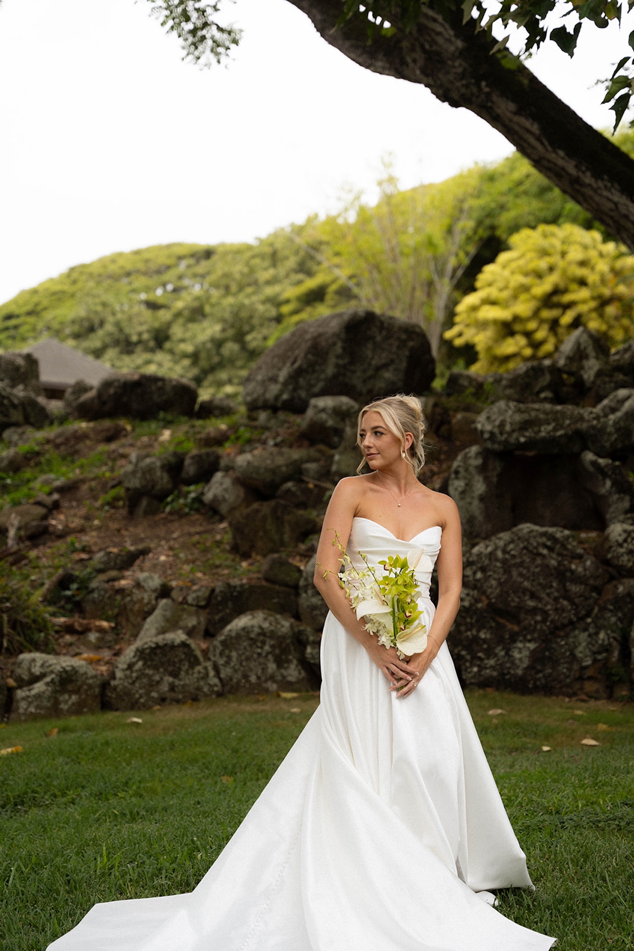 Bride standing solo against a backdrop of lava rocks and vibrant tropical trees, holding a white and green bouquet.