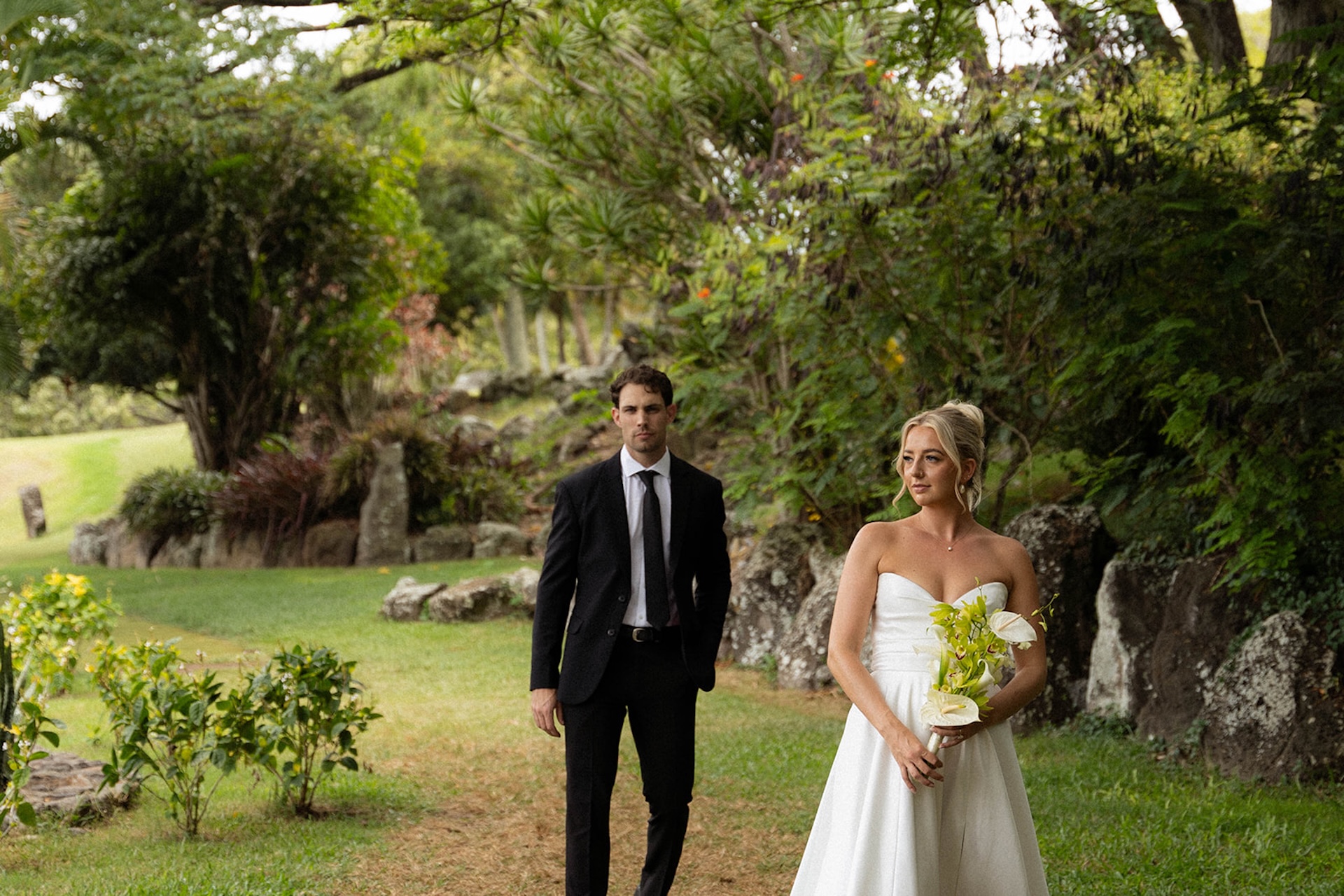 Bride holding a tropical bouquet while walking ahead of the groom through lush gardens filled with greenery and volcanic rock.