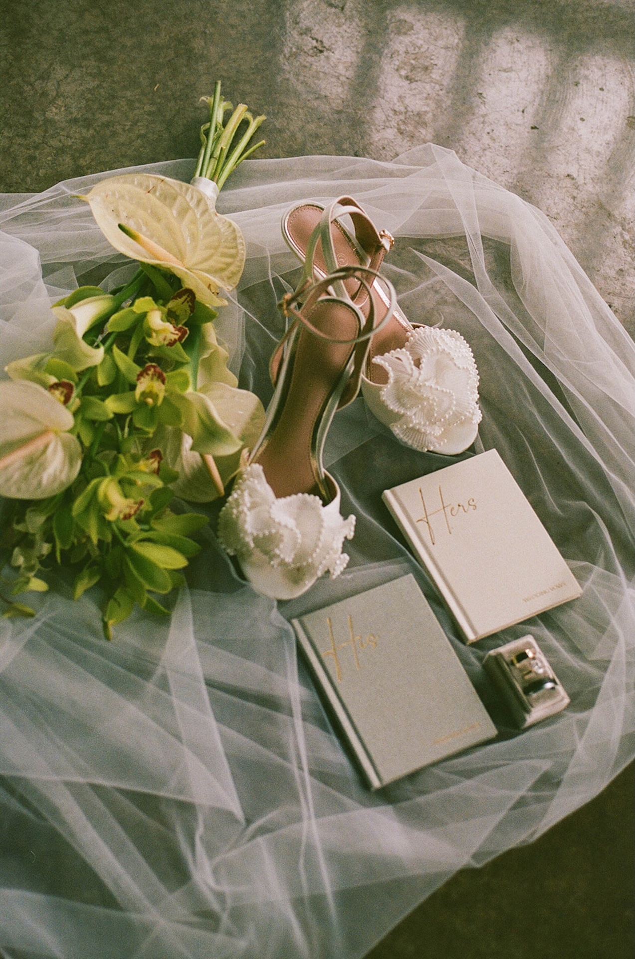 Flat Lay of white heels with orchids, His and Hers vows, the wedding rings, the brides orchid bouquet, laying on top of her veil.