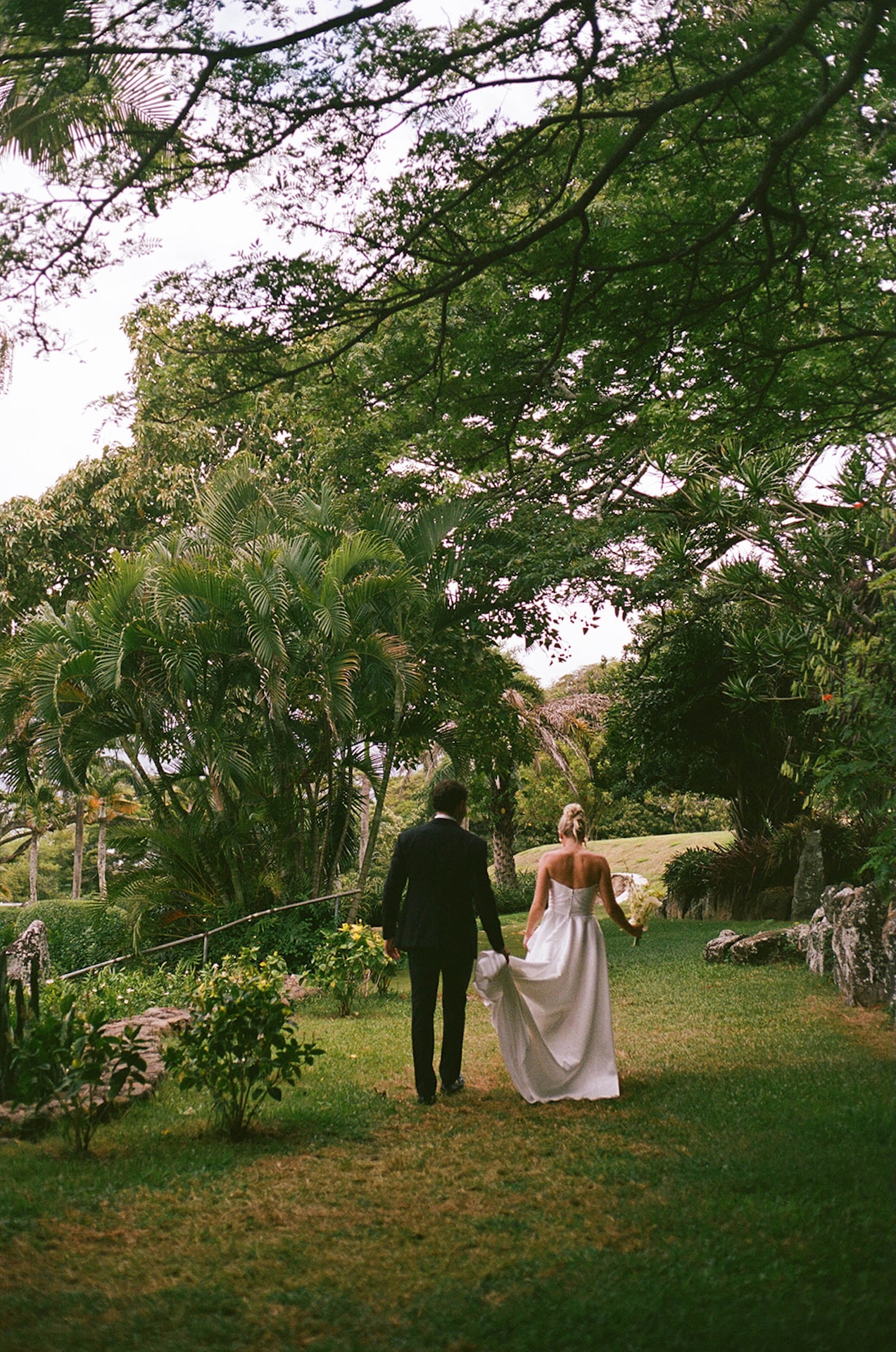 Film photo of the bride and groom walking hand in hand under lush trees at Kualoa Ranch for their hawaii wedding planning.