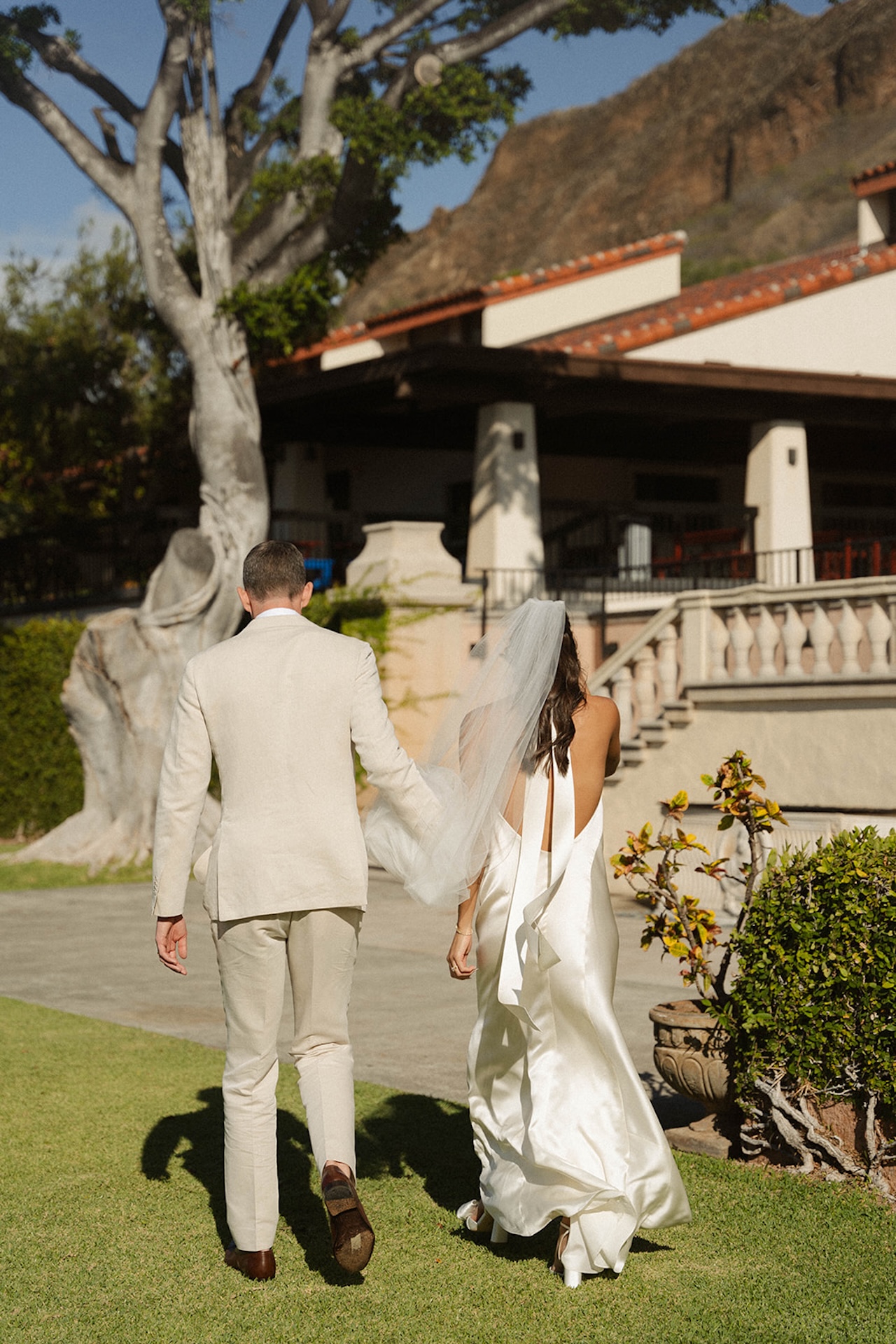 The couple walks hand in hand across the lawn, her silk dress trailing behind in the golden light, radiating intimate wedding elegance.