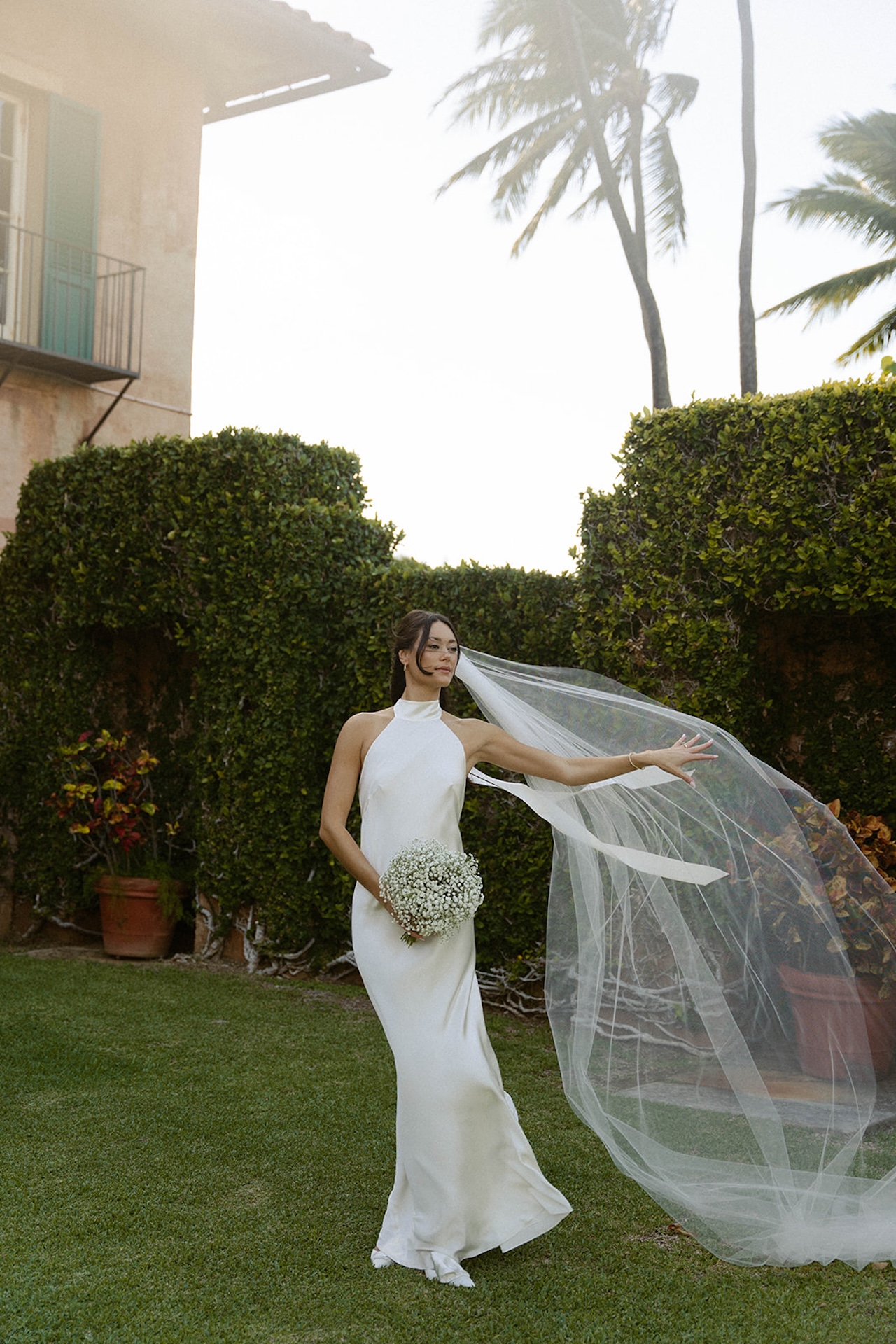 The bride stands elegantly in a halter satin gown, her veil flowing in the breeze against lush greenery — a scene from one of the best destination wedding locations.