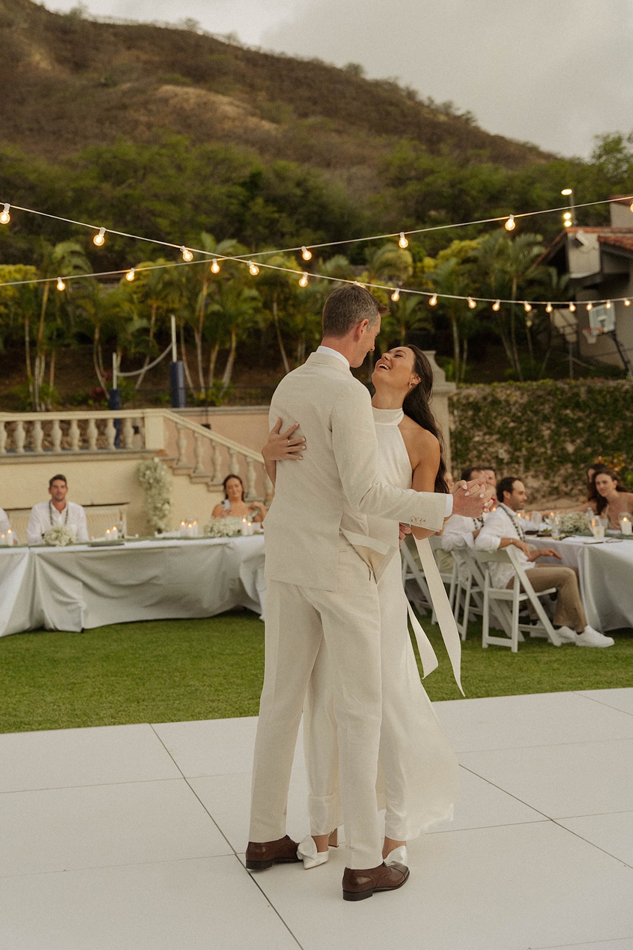 The couple shares a romantic first dance under string lights and mountain views, capturing the essence of an intimate wedding celebration outdoors.