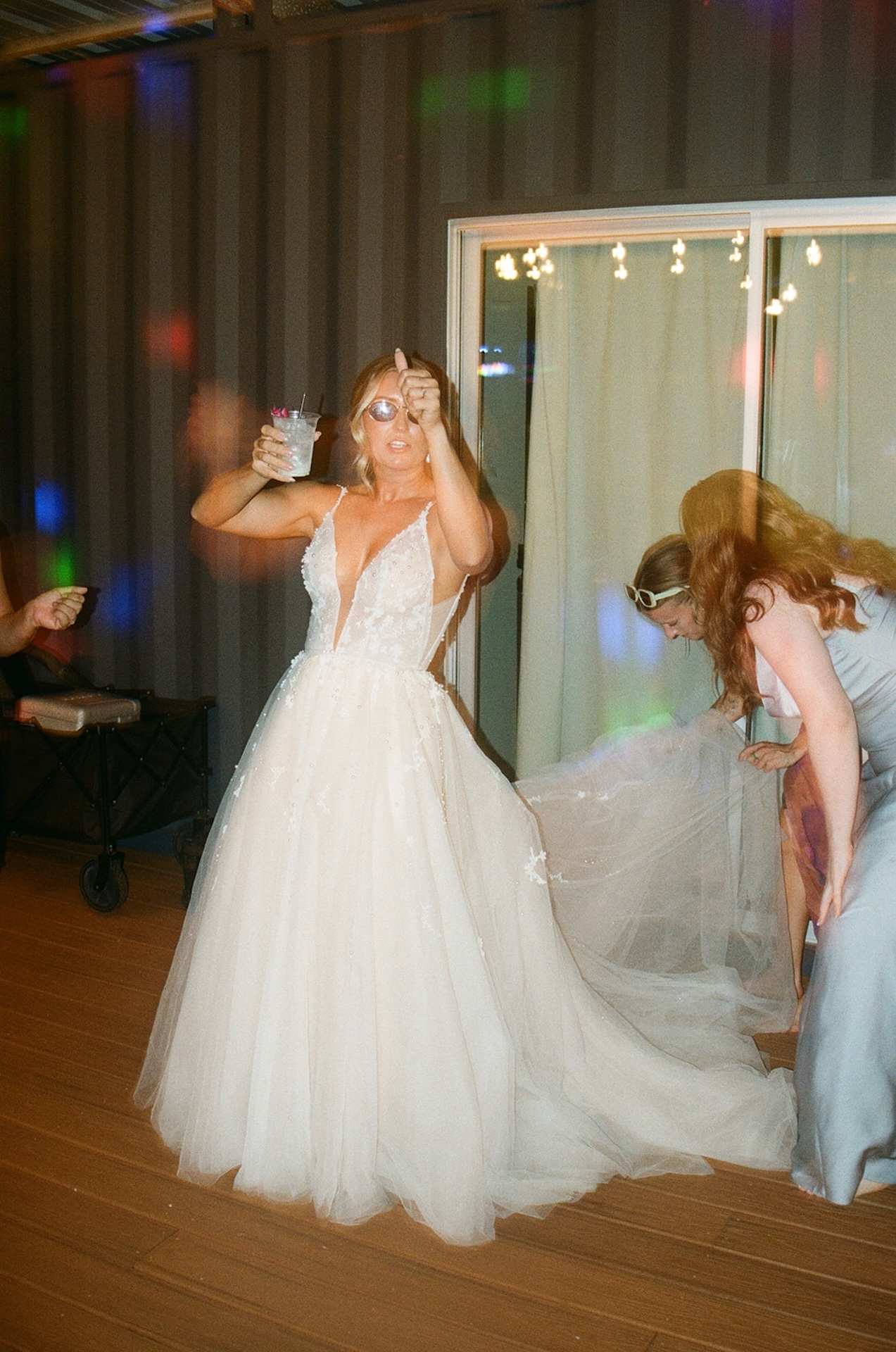 Bride dancing with friends in her wedding dress, holding a drink and smiling under the lights.