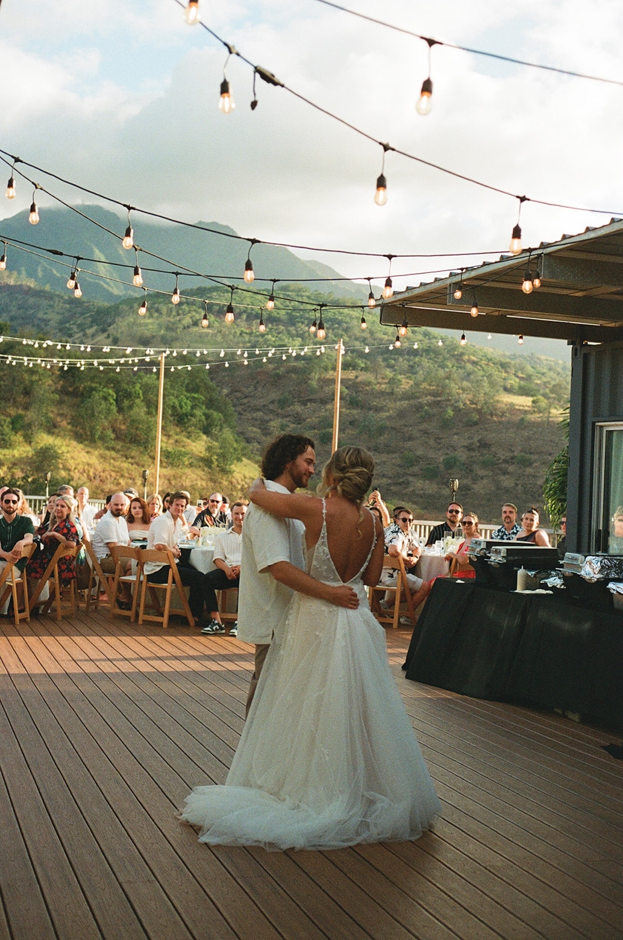 Couple sharing their first dance as friends and family watch, surrounded by string lights and scenic mountain views.