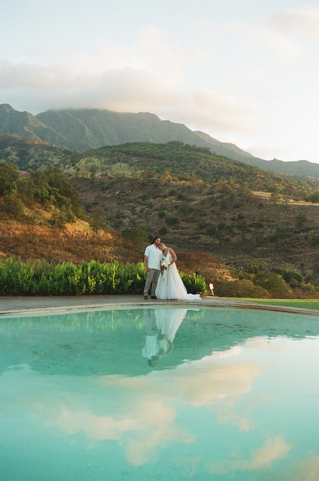 Bride and groom embracing beside a reflective pool as golden sunlight hits the mountain range behind them.