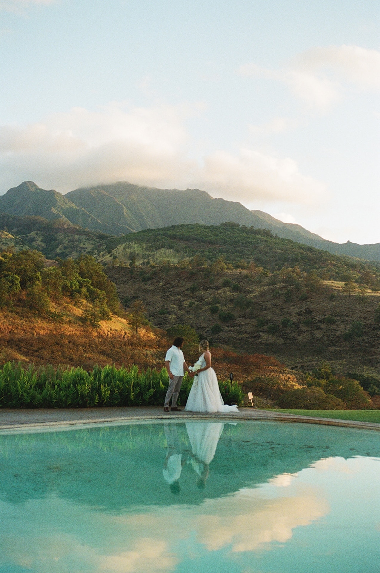 Couple holding hands at sunset near the water, surrounded by greenery and the peaceful beauty of Wedding Venues in Hawaii.